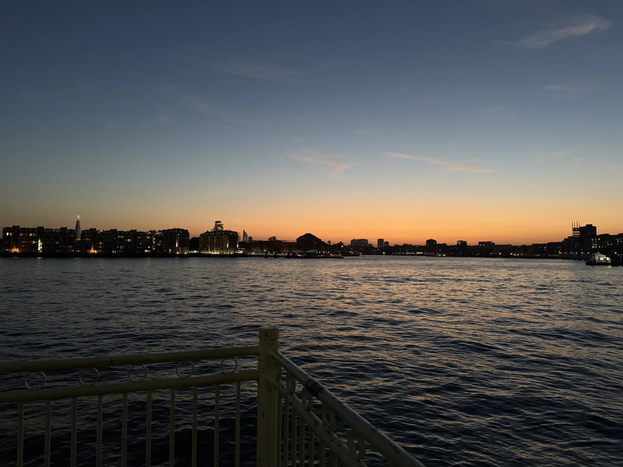 View of a city skyline at sunset with the sky transitioning from orange near the horizon to deep blue at the top, seen from the water with reflections on the surface. Buildings are silhouetted in the distance, and part of a boat or dock rail is visib