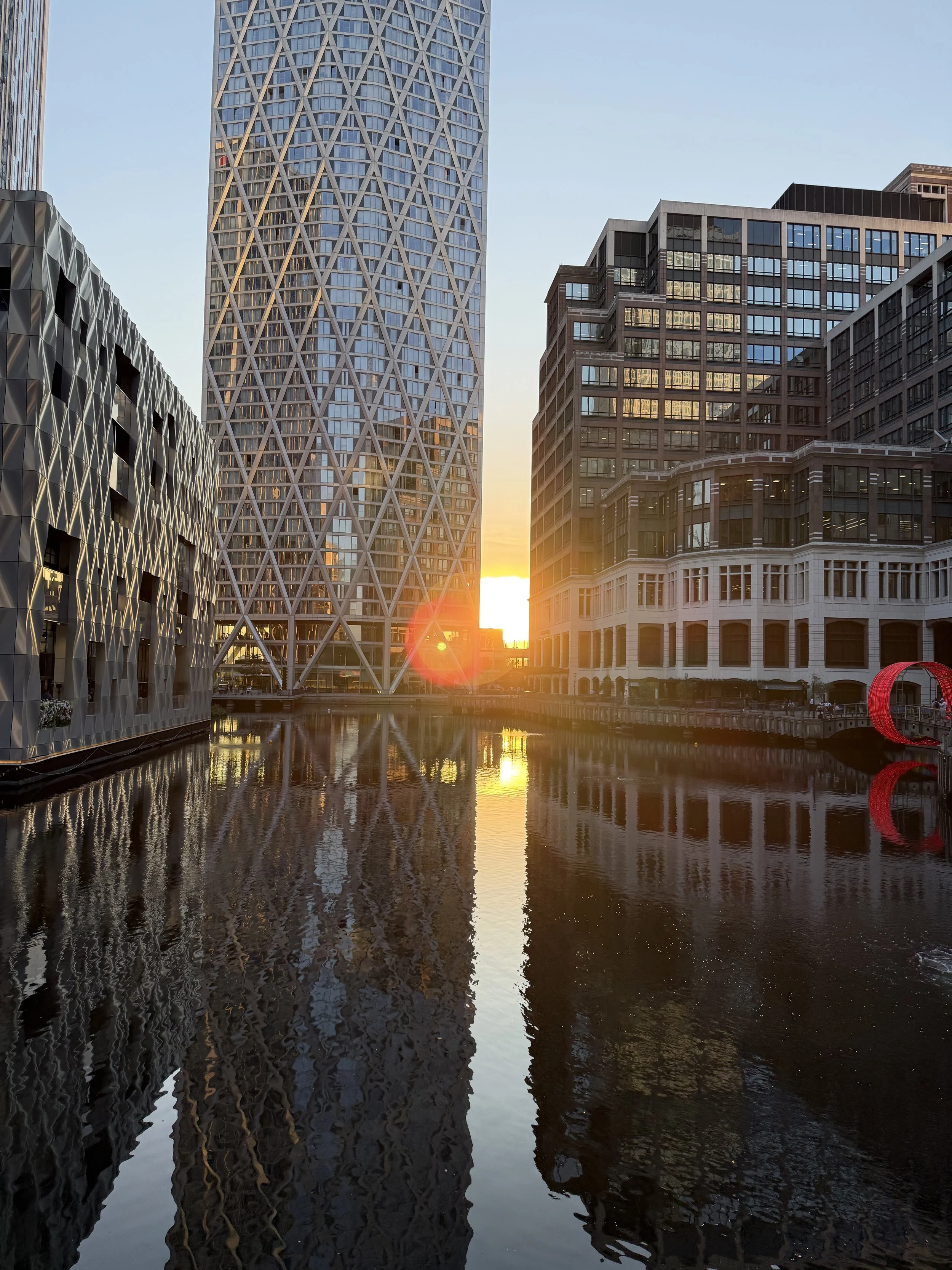 Tall modern skyscrapers reflecting in a river at sunset.