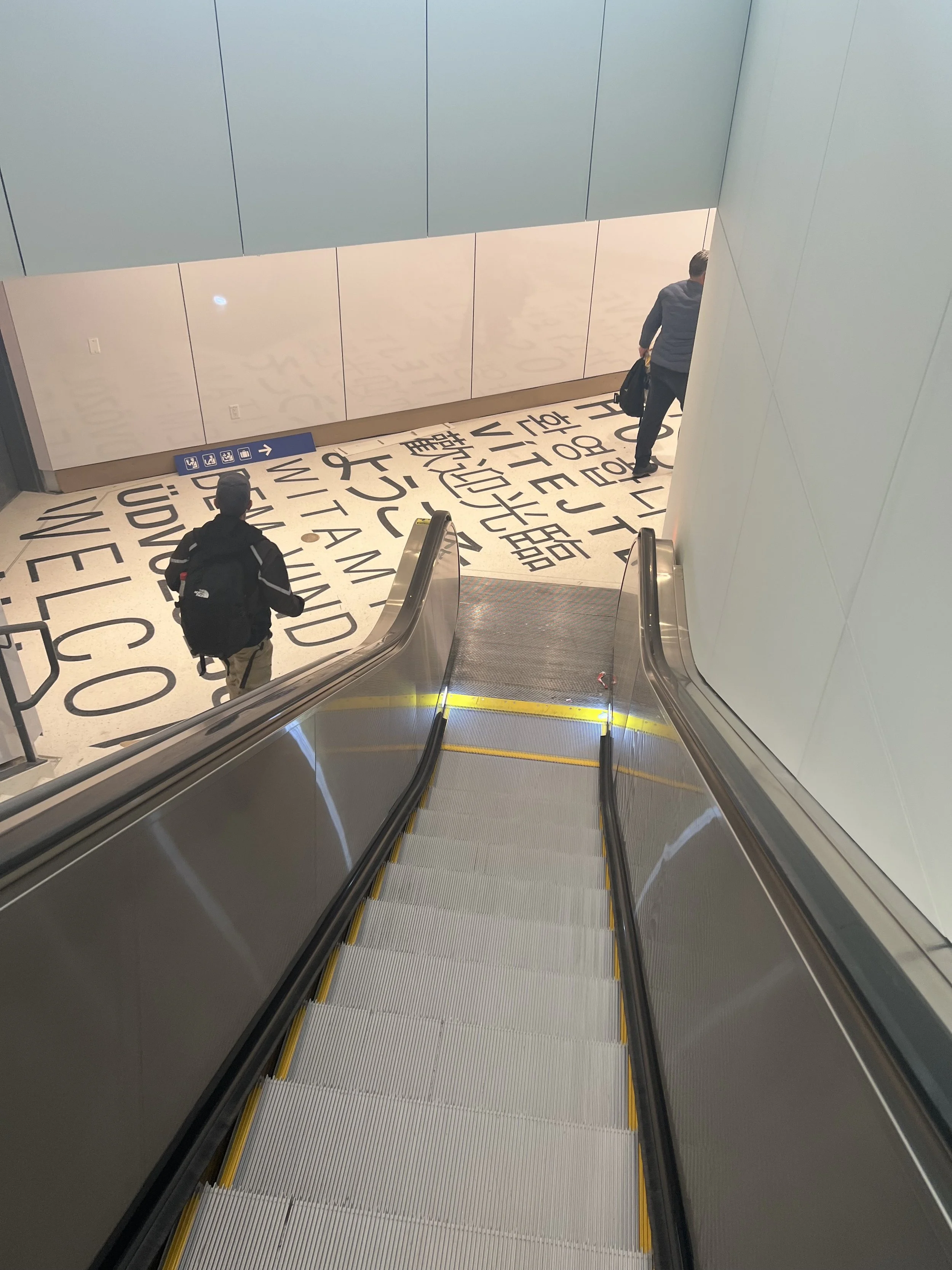 An escalator descending in an indoor location, with two people on it. One person is sitting on the escalator wearing a black jacket and backpack, and the other is standing at the top holding a black bag. The floor features large black letters and wor