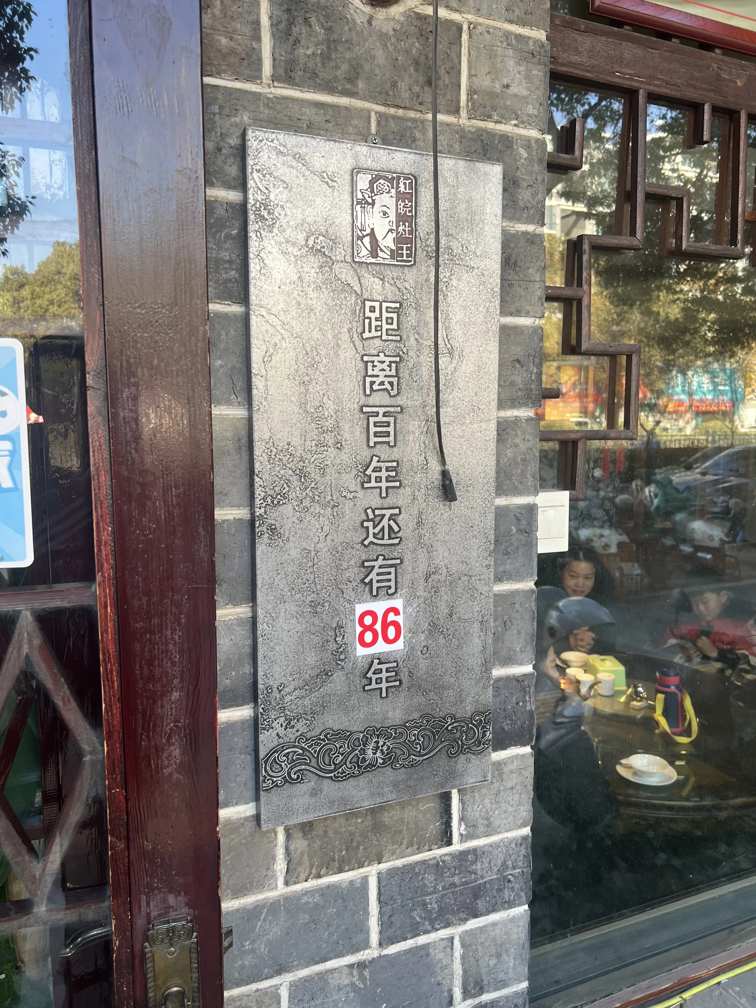 A sign on a brick wall with Chinese characters and a graphic of a woman’s face, with reflections of people and trees in the glass window showing a dining scene inside a restaurant.