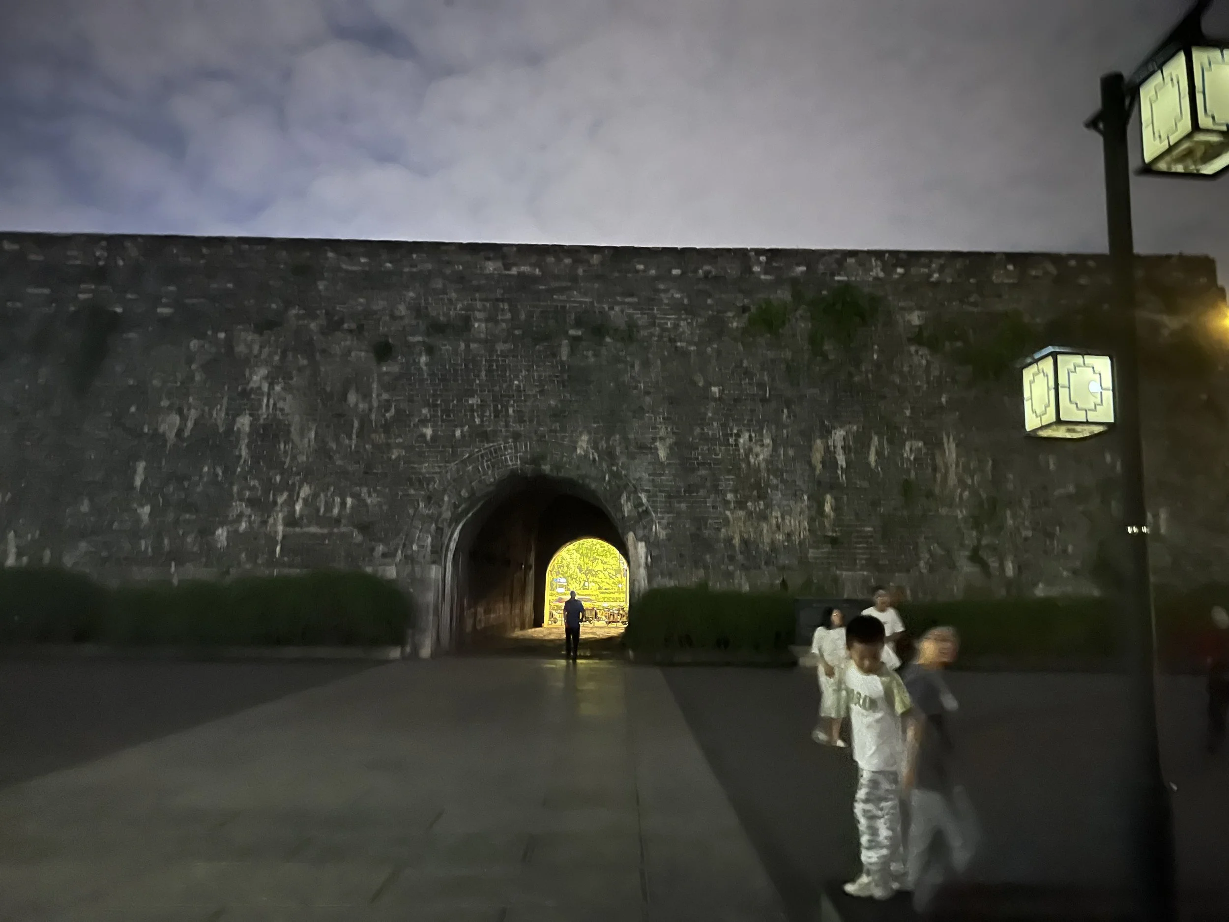 Night scene with stone archway tunnel illuminated by yellow-green lights, people walking, and a bright yellow gate visible in the distance.