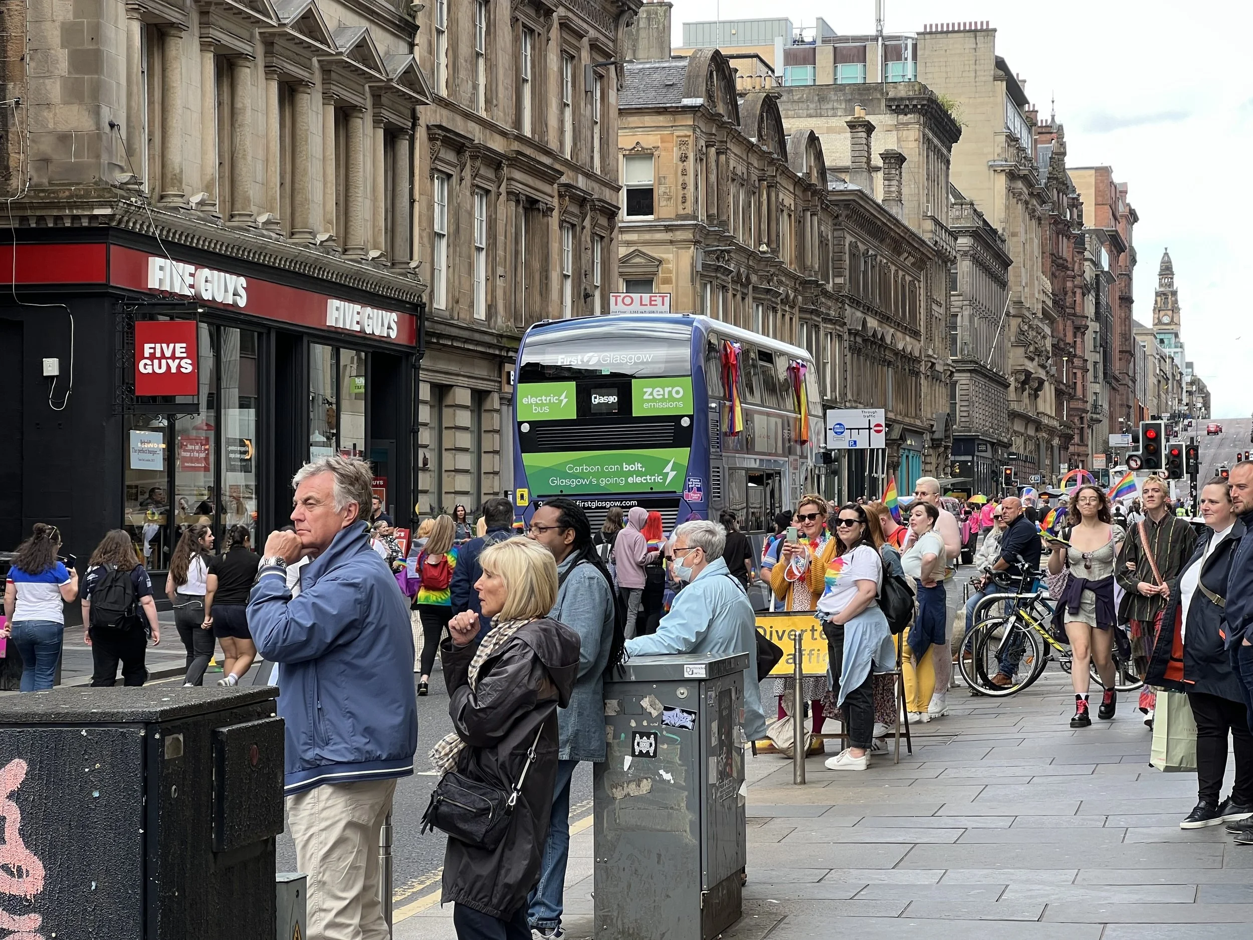 Street scene in Glasgow with a large crowd of people, some holding rainbow flags, gathered for a pride event. There are historic buildings, a bus promoting electric transportation, and storefronts including a Five Guys restaurant, with some people wa