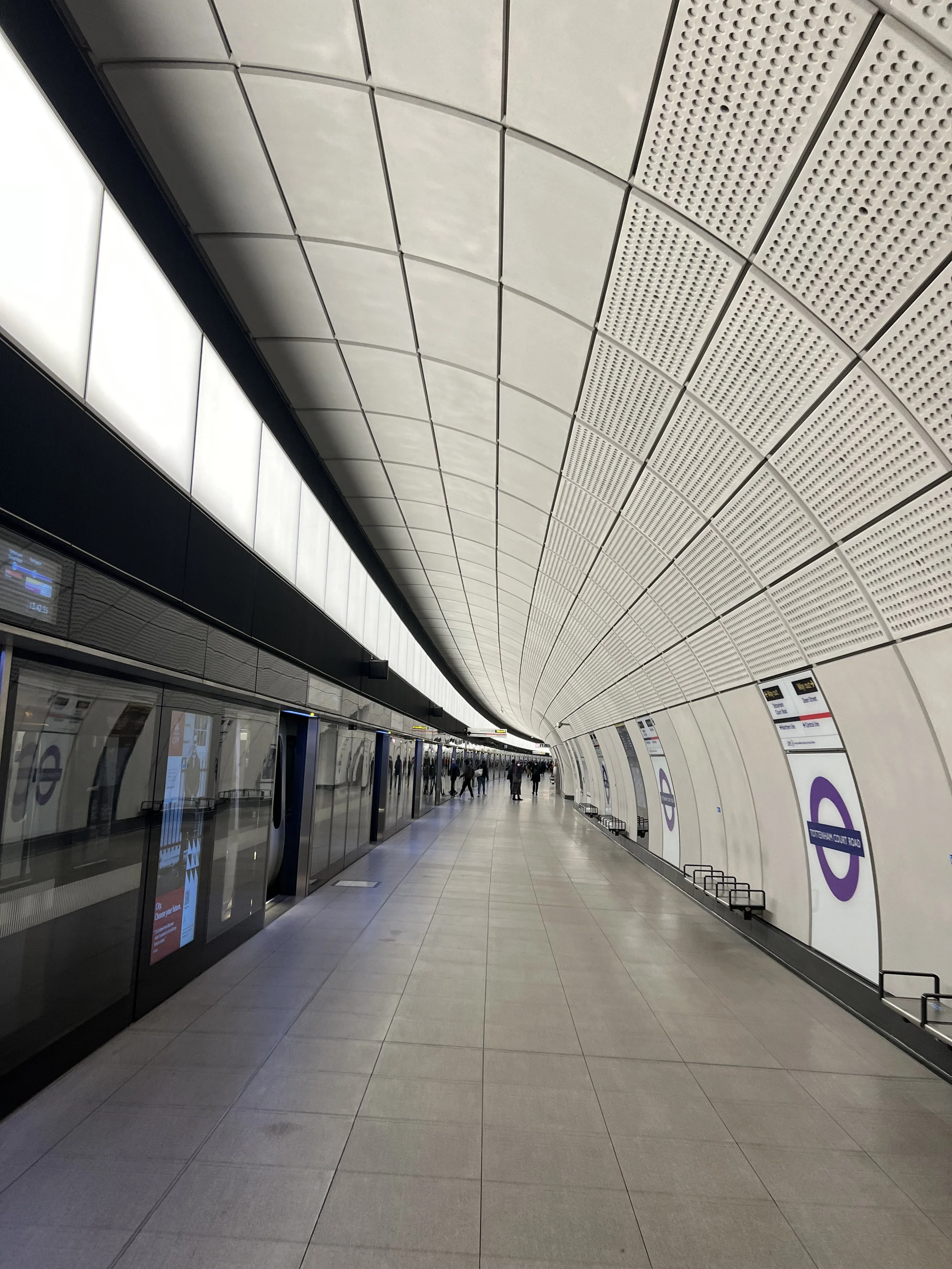 Indoor view of a modern train station platform with a curved ceiling, glass doors, digital information screens, and plenty of people walking.