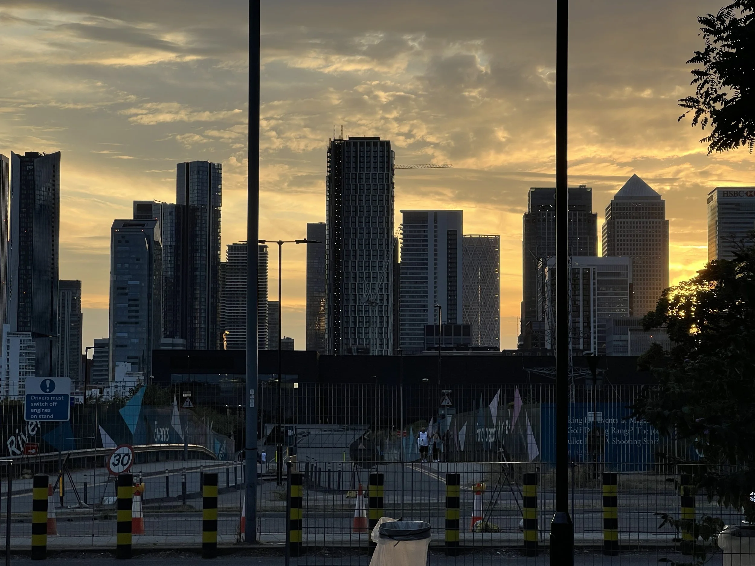 City skyline with tall buildings at sunset, viewed through a fenced area with traffic cones and street signs in the foreground.