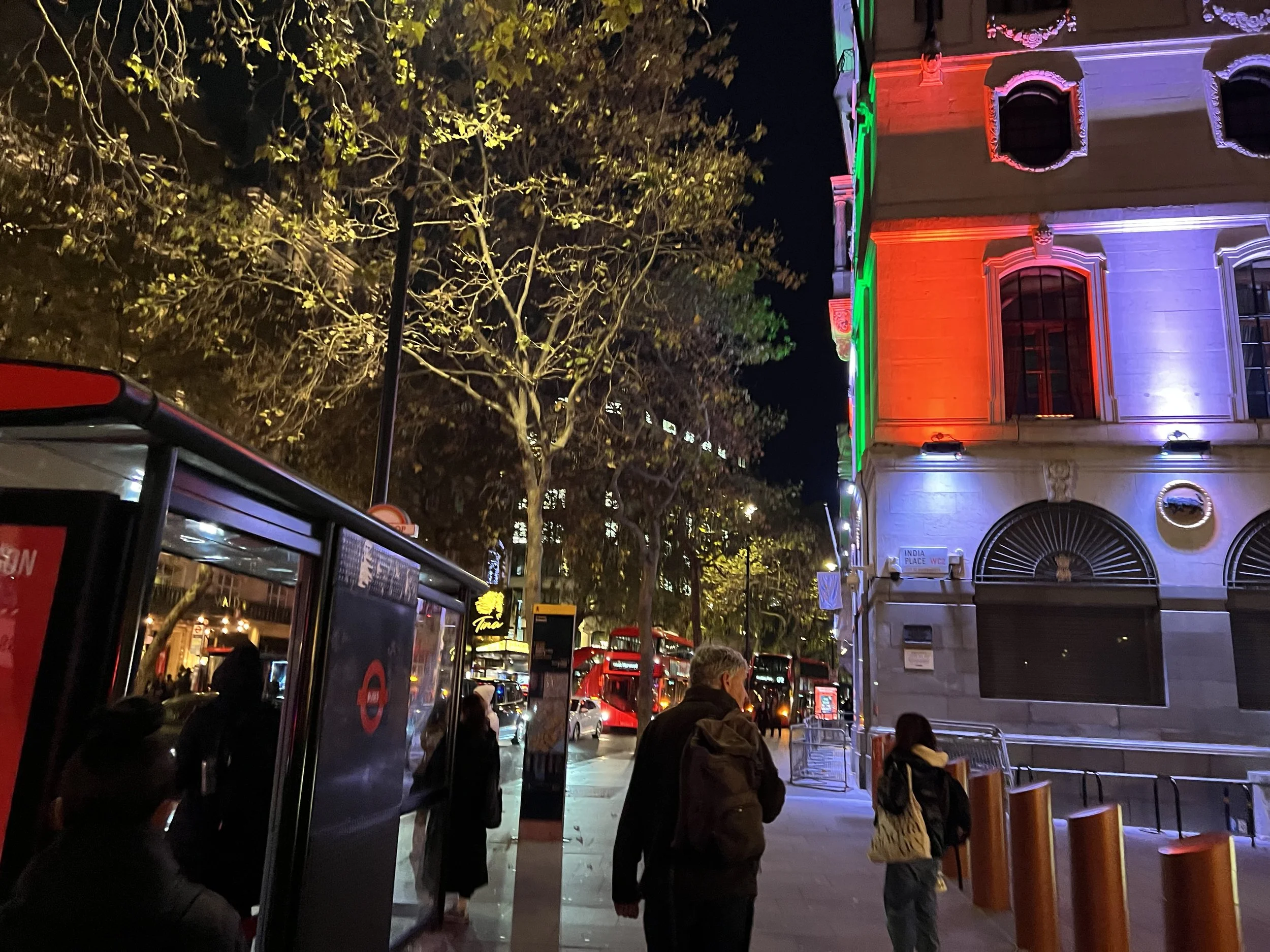 Nighttime street scene with people waiting at a bus stop, illuminated building decorated with colorful lights, trees, and parked cars.