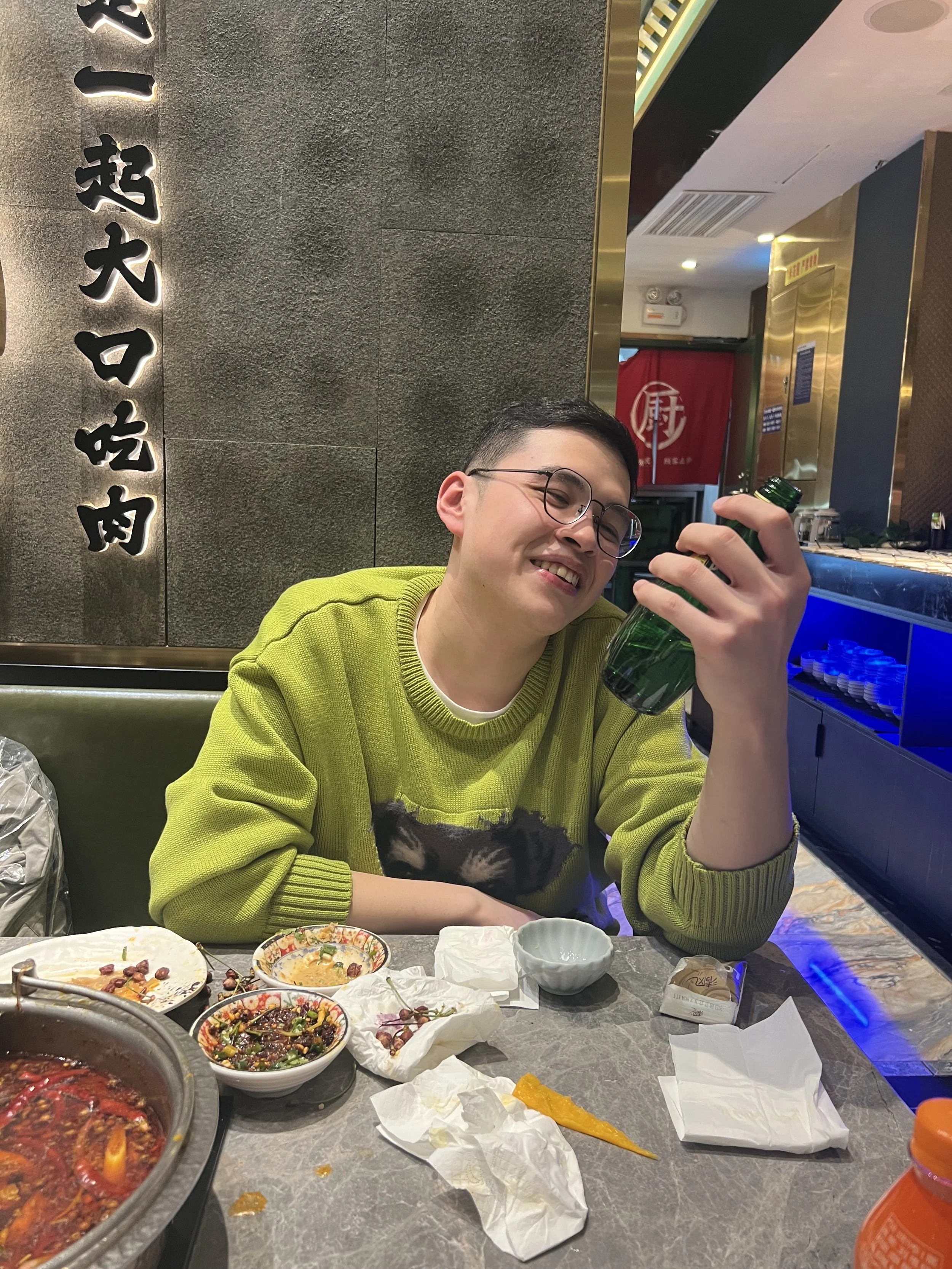 A young man with glasses smiling and holding a green bottle at a dining table with leftover food bowls, crumpled napkins, and food containers, inside a restaurant with traditional Asian decor.