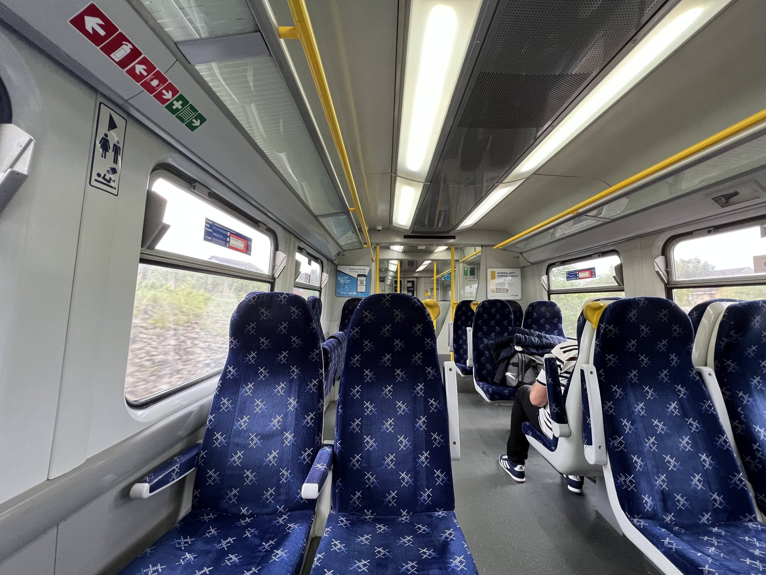 Interior of a train showing blue patterned seats, yellow handrails, and large windows, with a passenger standing near the back.