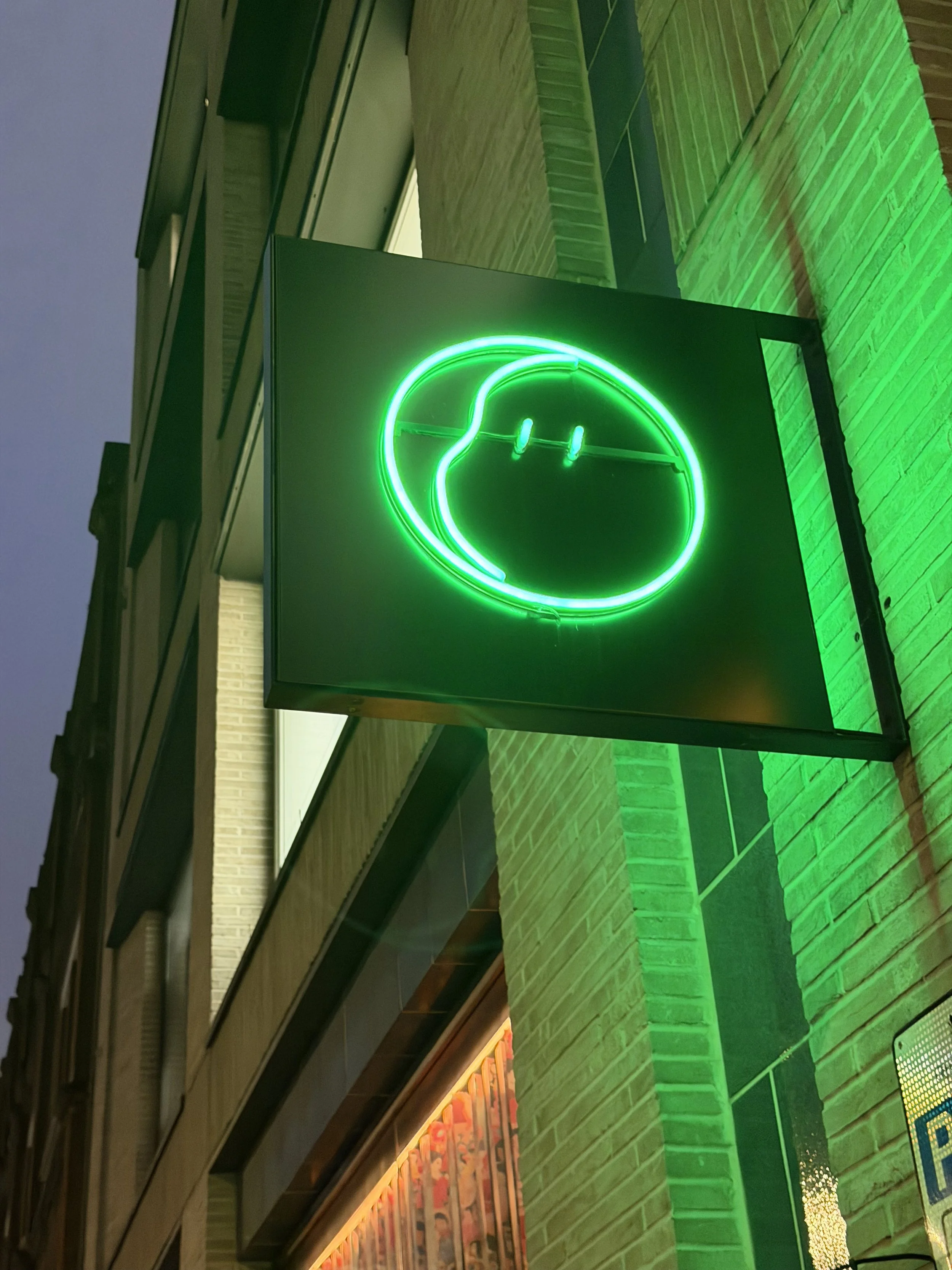 Green neon sign in the shape of a Japanese lucky cat with a raised paw, mounted on a building with brick and wood paneling, illuminated with green lights.
