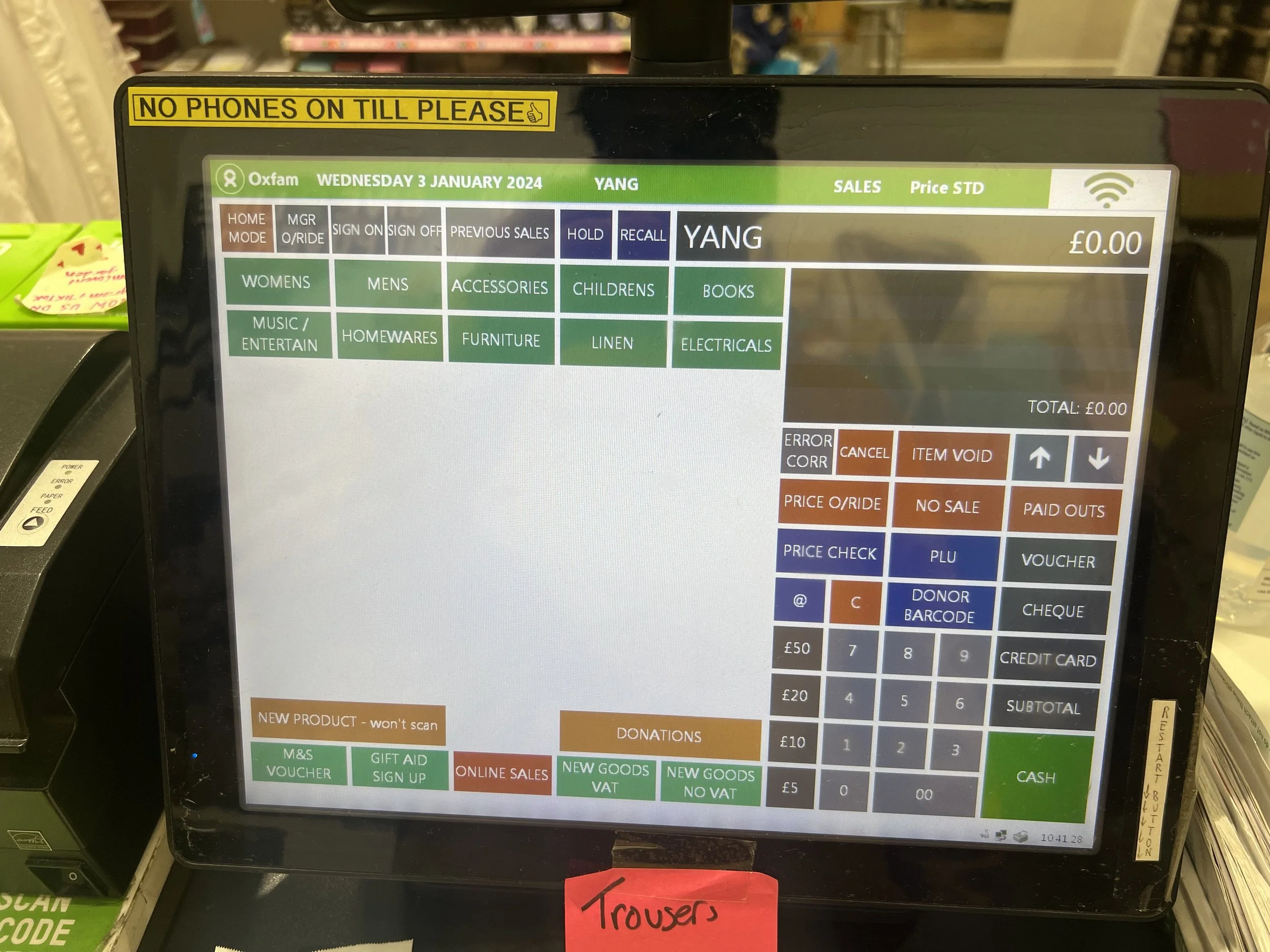 Close-up of a store checkout screen displaying various buttons and options, with a yellow sign above it that reads 'NO PHONES ON TILL PLEASE.' The screen shows the date Wednesday, 3 January 2024, and the name 'YANG.' The screen is part of a checkout 