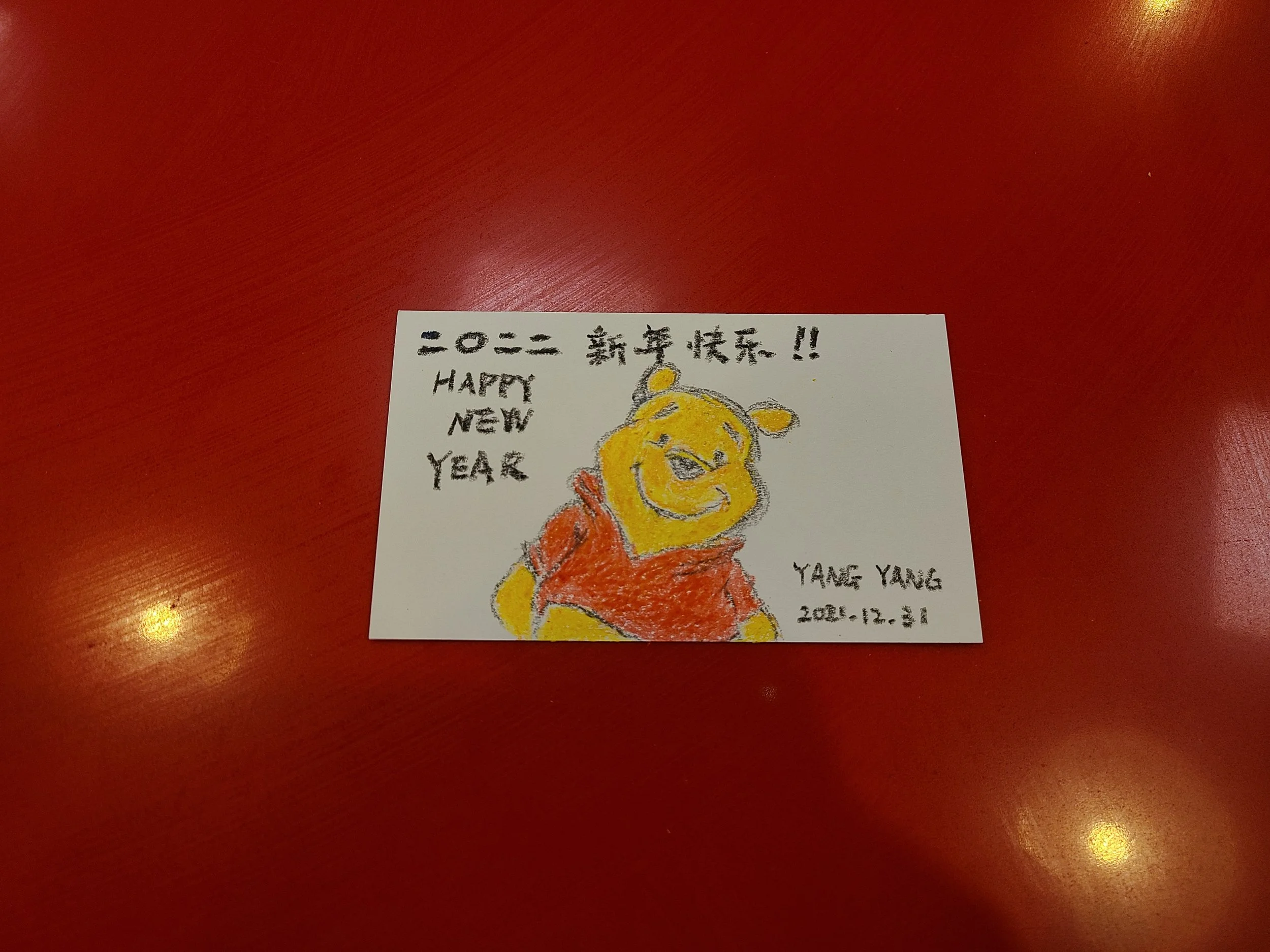 A colorful hand-drawn card with a yellow bear mascot wearing red clothing, celebrating New Year 2022, with handwritten messages in Japanese and English, placed on a red surface.