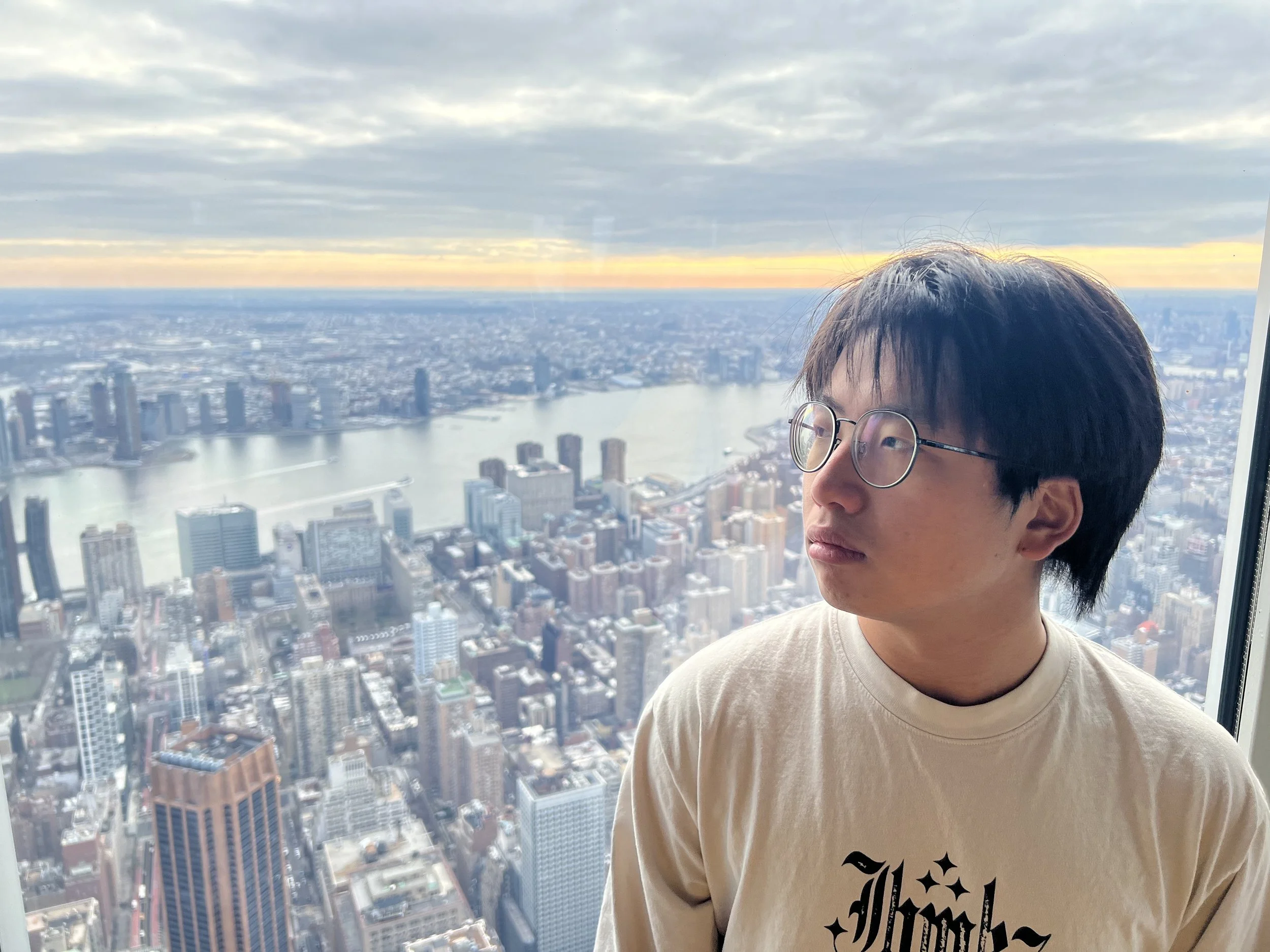 A young man wearing glasses and a beige T-shirt with black text, looking to the right, with a city skyline and river visible in the background from a high vantage point.