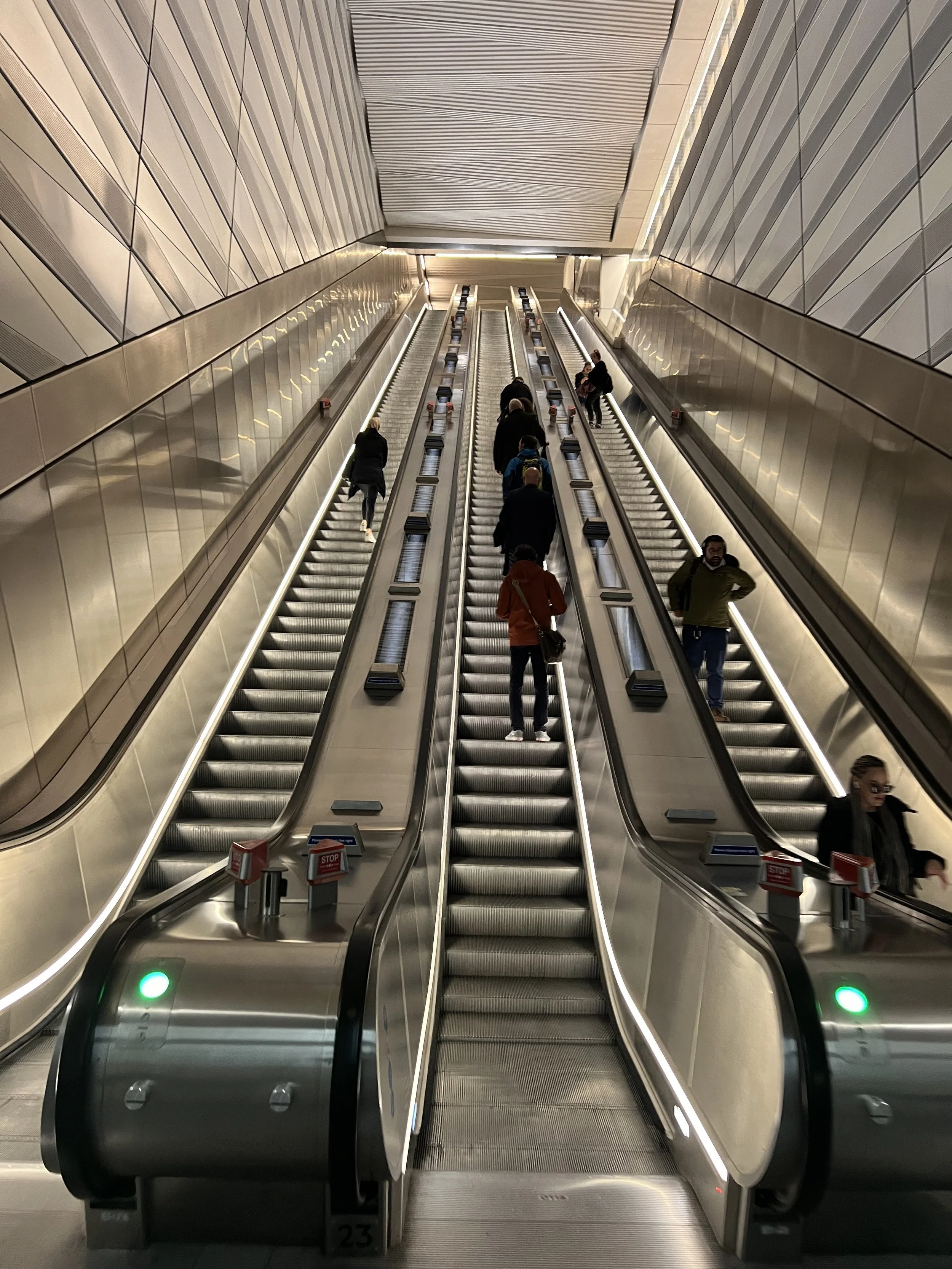 Inside an airport, two escalators with several passengers riding up and down, surrounded by metallic walls and panels.