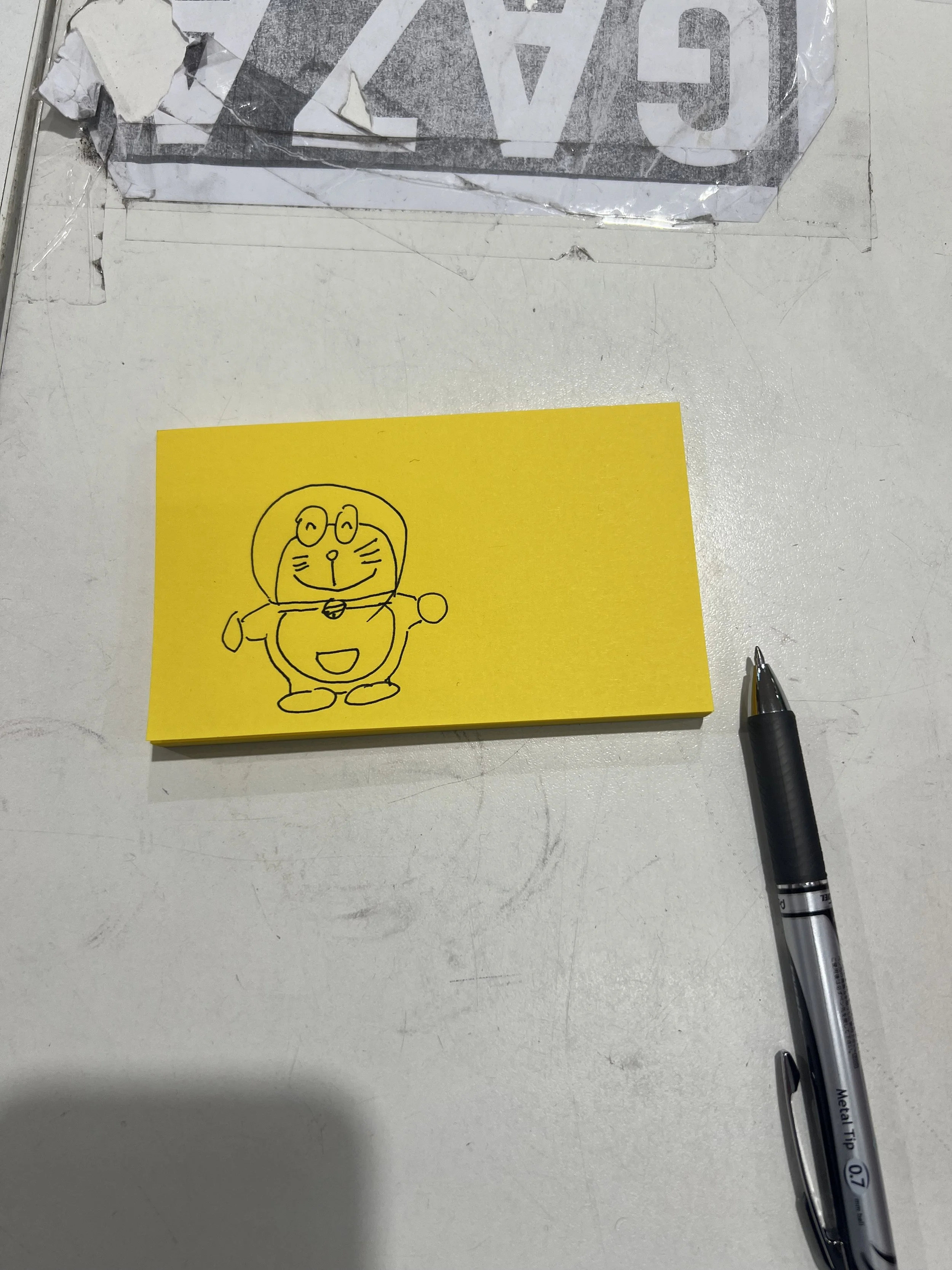 A yellow sticky note with a simple black line drawing of a cartoon cat wearing glasses and a collar, placed on a white surface. To the right of the note is a silver ballpoint pen with a black grip.