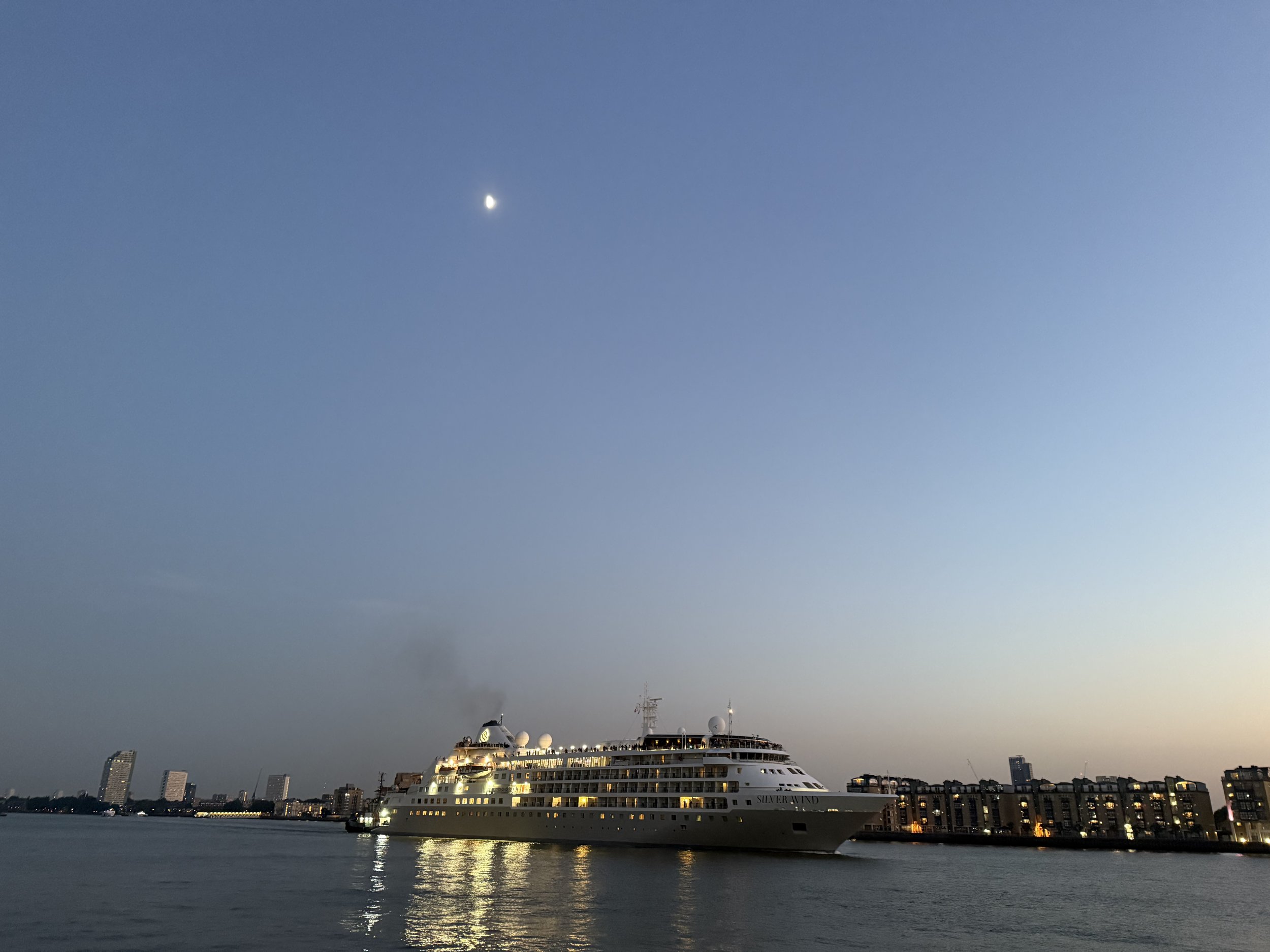 Large white cruise ship named Silver Wind docked at a city waterfront during dusk, with buildings and a clear sky with the moon overhead, calm water reflecting the ship and city lights.