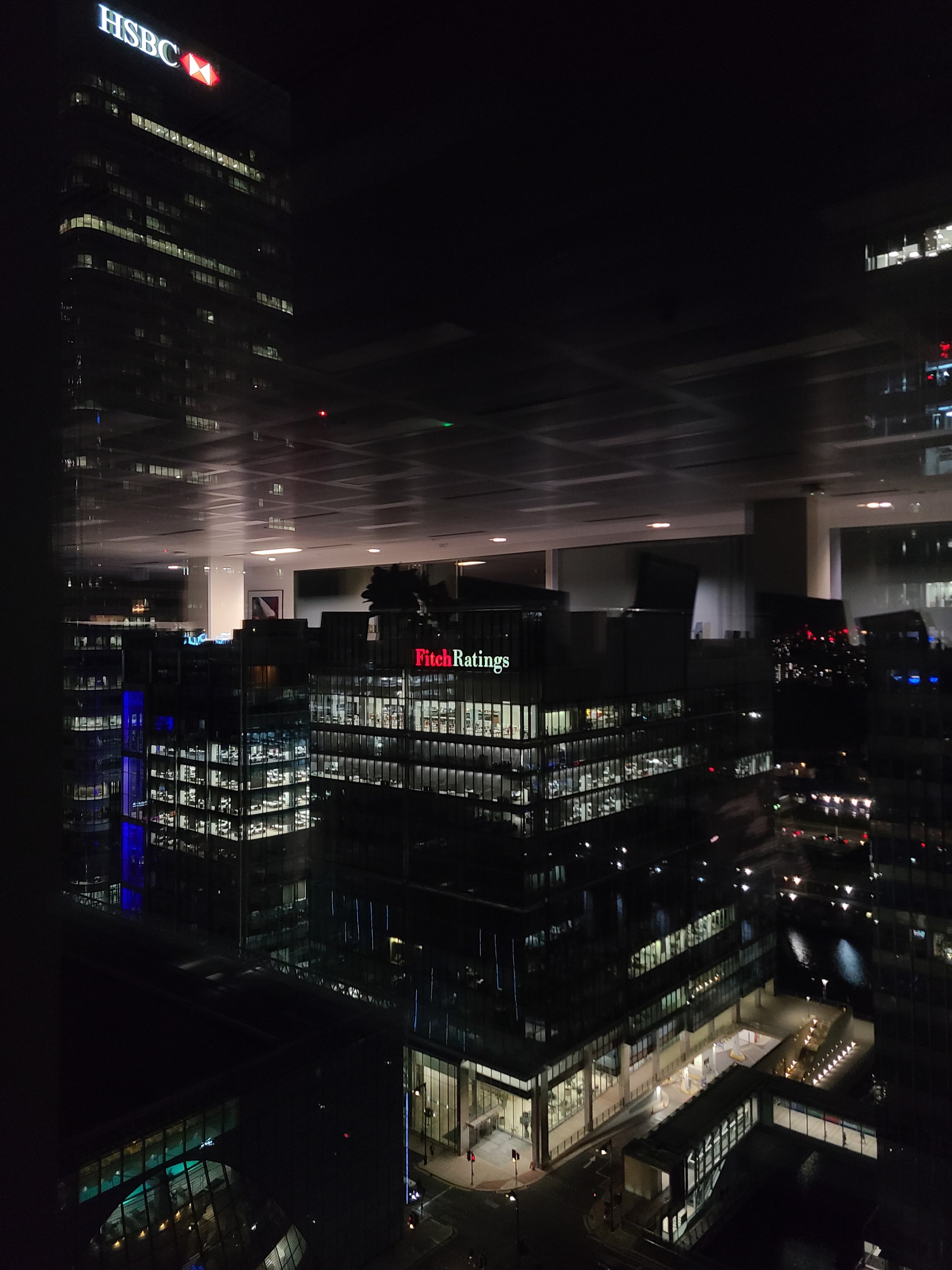 Nighttime cityscape showing illuminated office buildings, including HSBC and Fitch Ratings, with office lights on and street-level activity.
