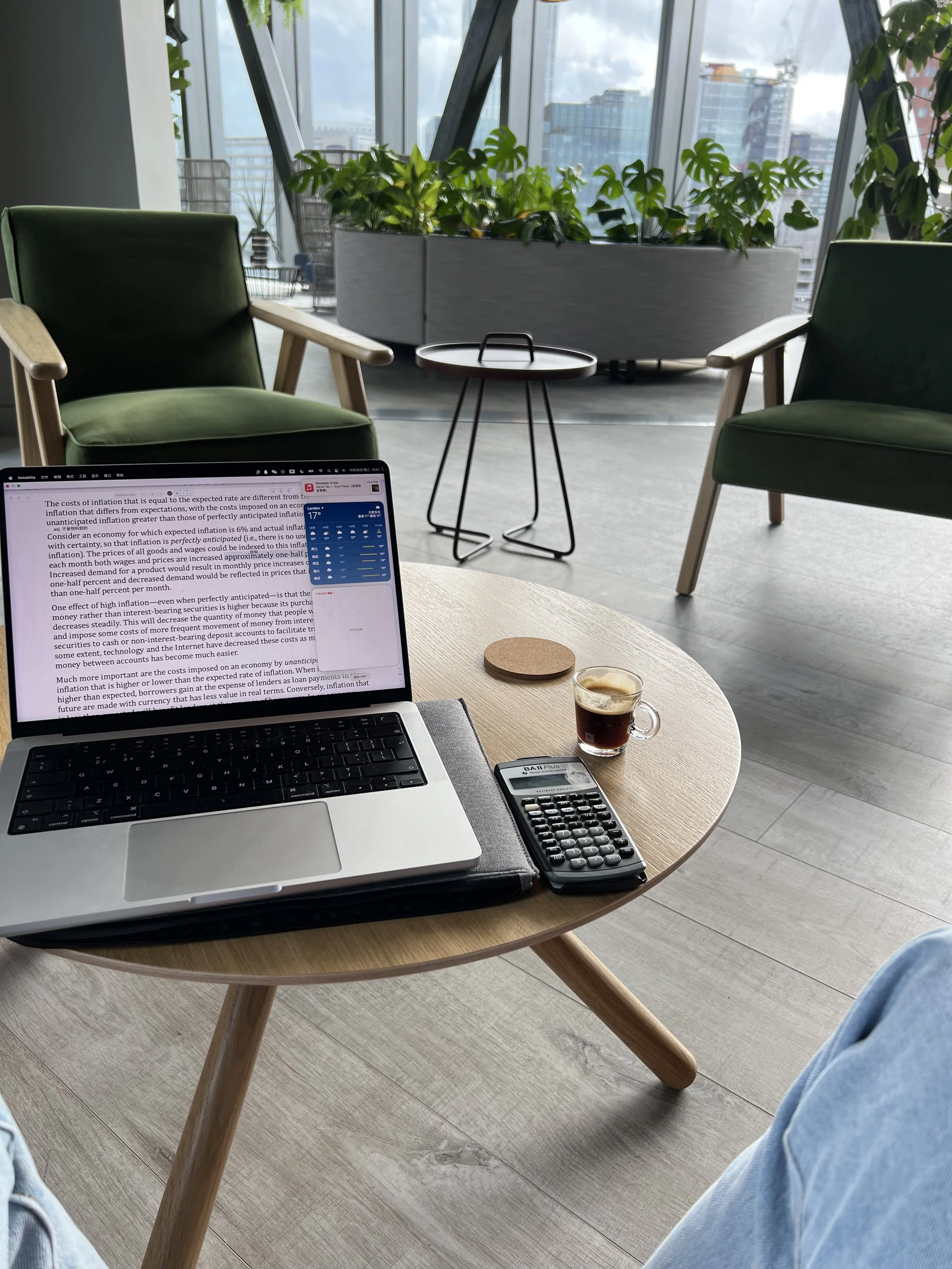 Indoor area with a wooden table, green armchairs, potted plants, large windows showcasing a cityscape, a laptop with reading material, a calculator, a coffee cup, and part of a person's denim-clad legs.