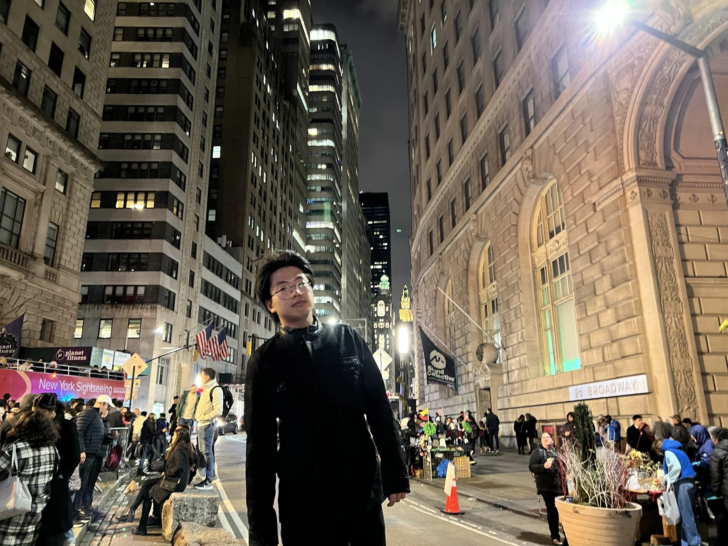 A woman with glasses and a black jacket stands in front of a busy street scene at night in New York City. The street is filled with people, vendor booths, and American flags. Tall buildings with illuminated windows surround the scene, with one buildi