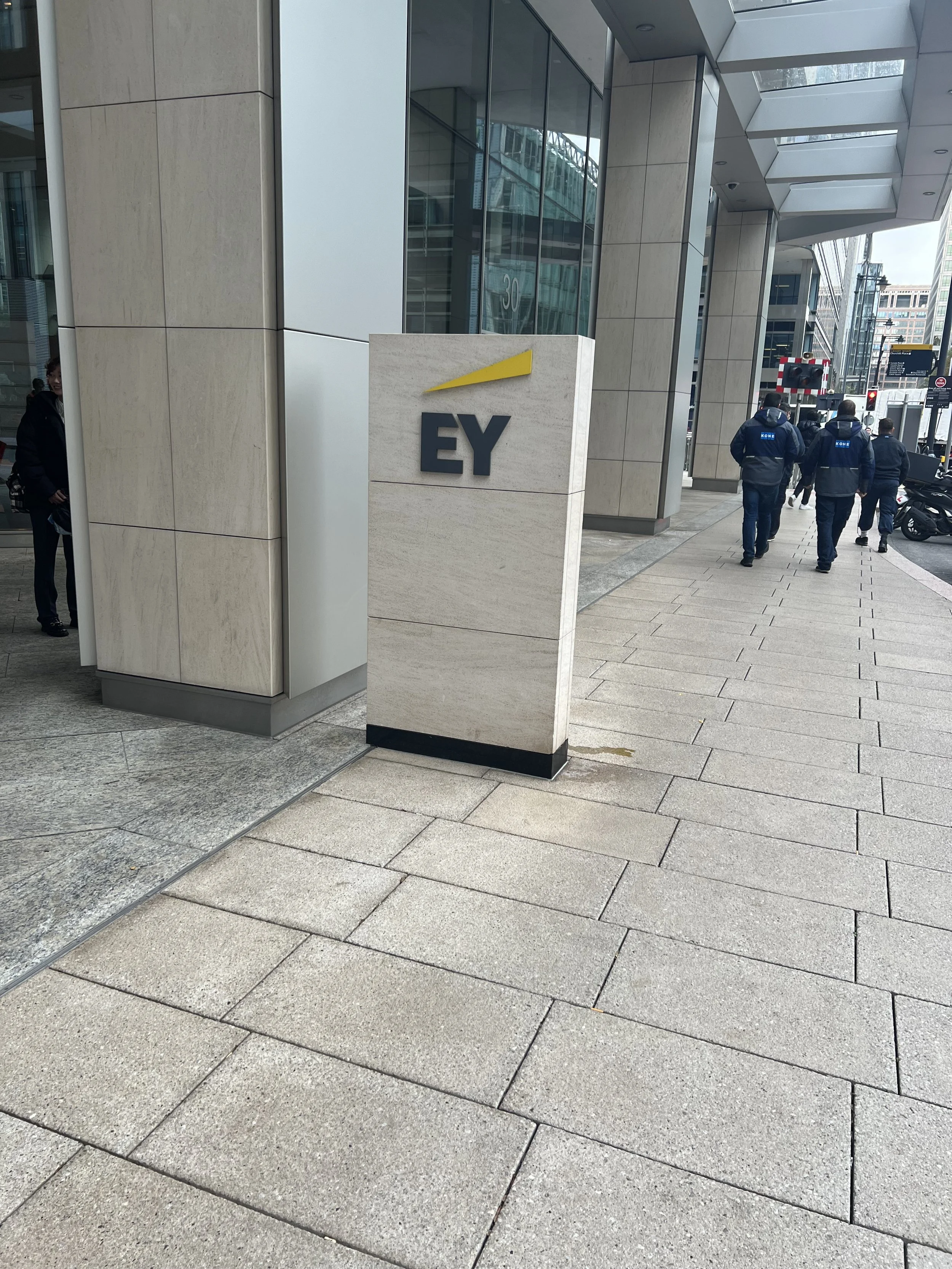 Exterior view of a modern office building with a sign for EY (Ernst & Young) on a stone column, with a sidewalk, people walking, and glass windows reflecting the street scene.