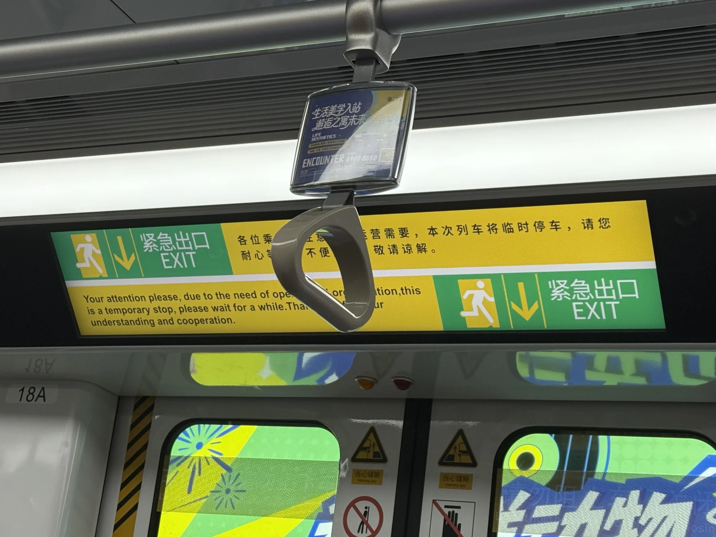 A bus interior showing a hanging sign with Chinese characters and English text that reads 'Lifestyle Aesthetics Encounter' along with a phone number. Below are various safety and informational signs, including a green emergency exit sign, caution sym