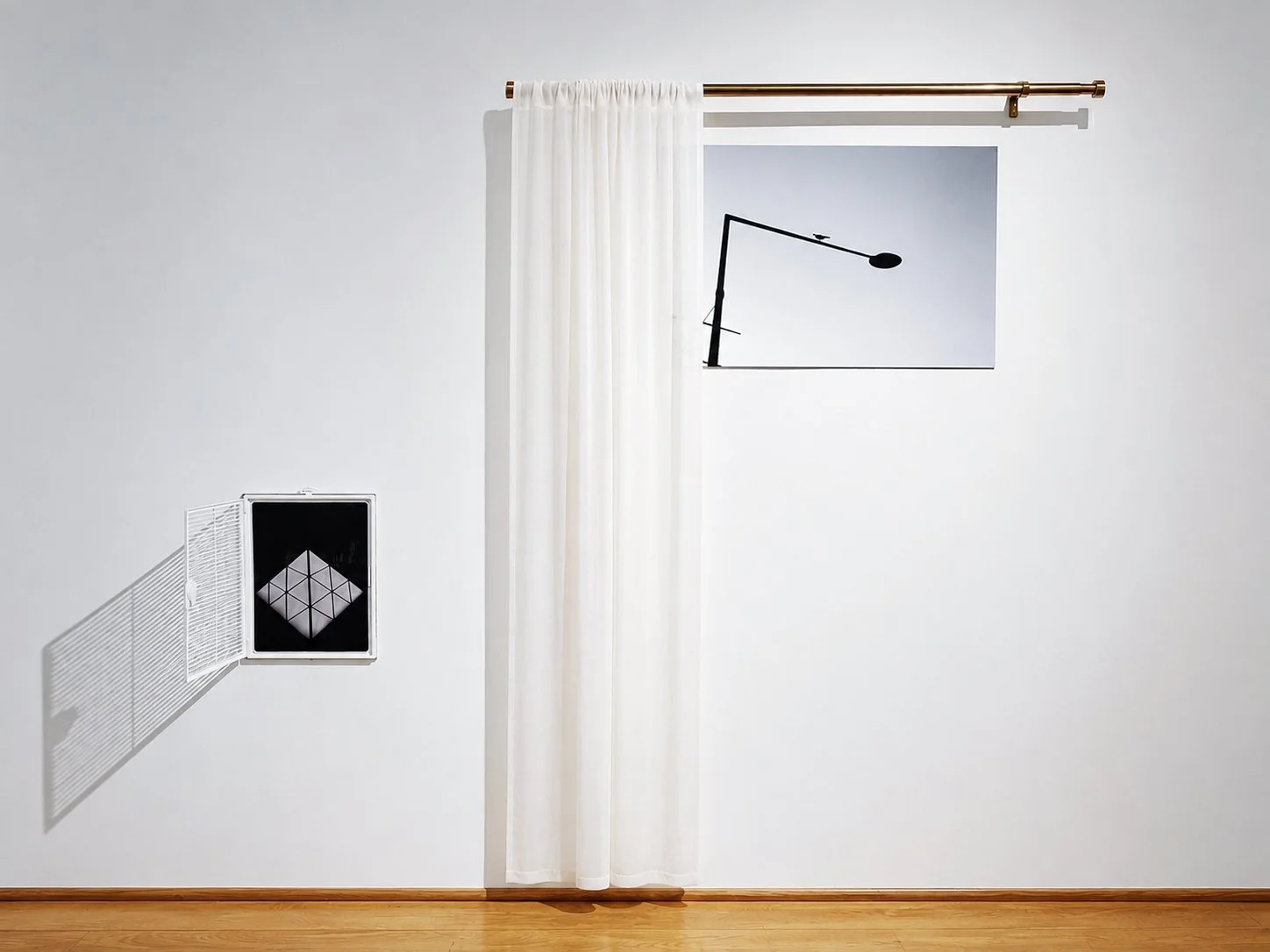 Minimalist white wall with a sheer white curtain hanging from a gold curtain rod, a framed poster of a black desk lamp, and an open small cabinet with geometric design elements casting a shadow on the wall.
