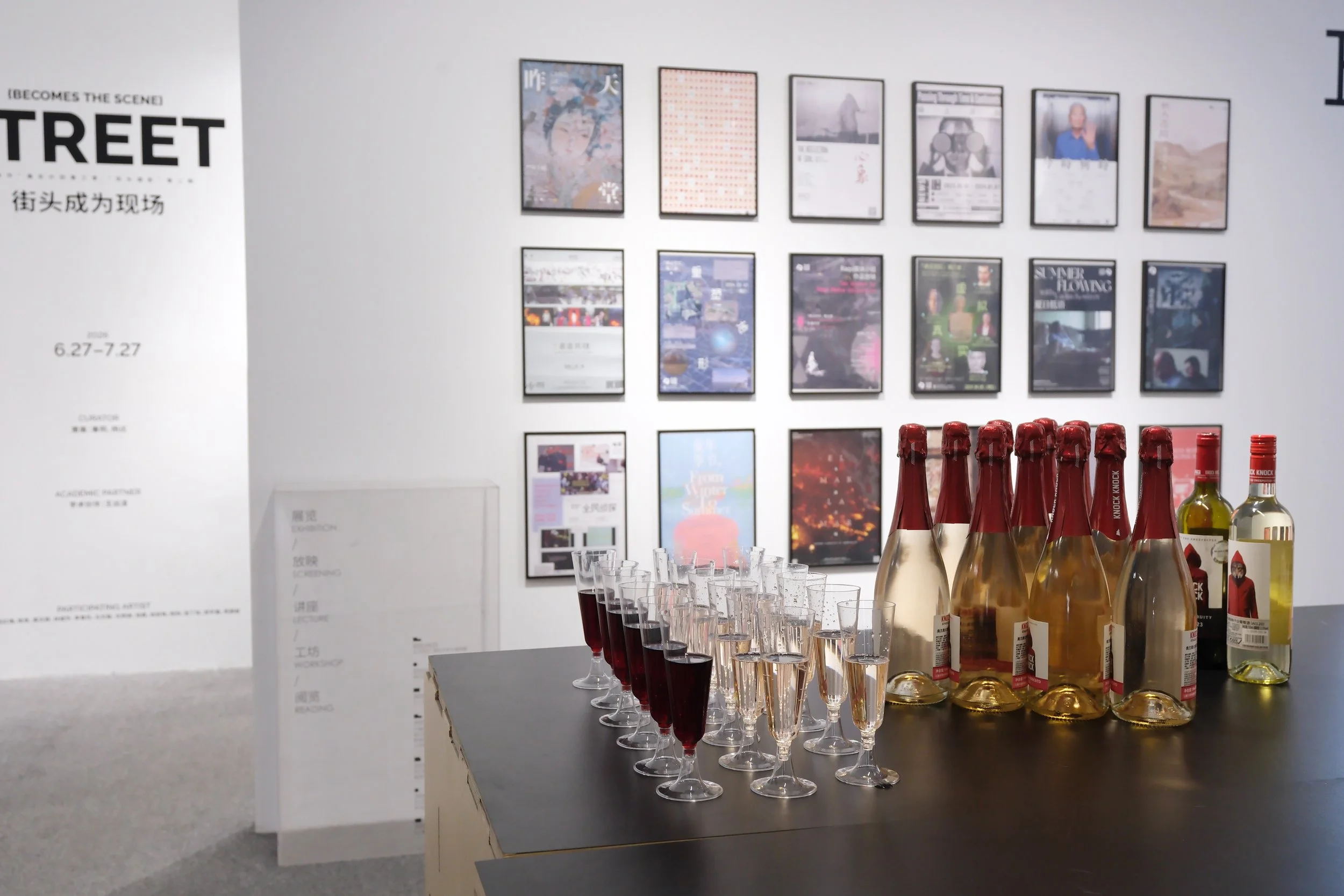 A display of bottles, likely of sparkling wine or champagne, arranged on a table alongside rows of filled glasses with red and clear liquids. In the background, framed posters and artwork are mounted on the wall of an art gallery or exhibition space.