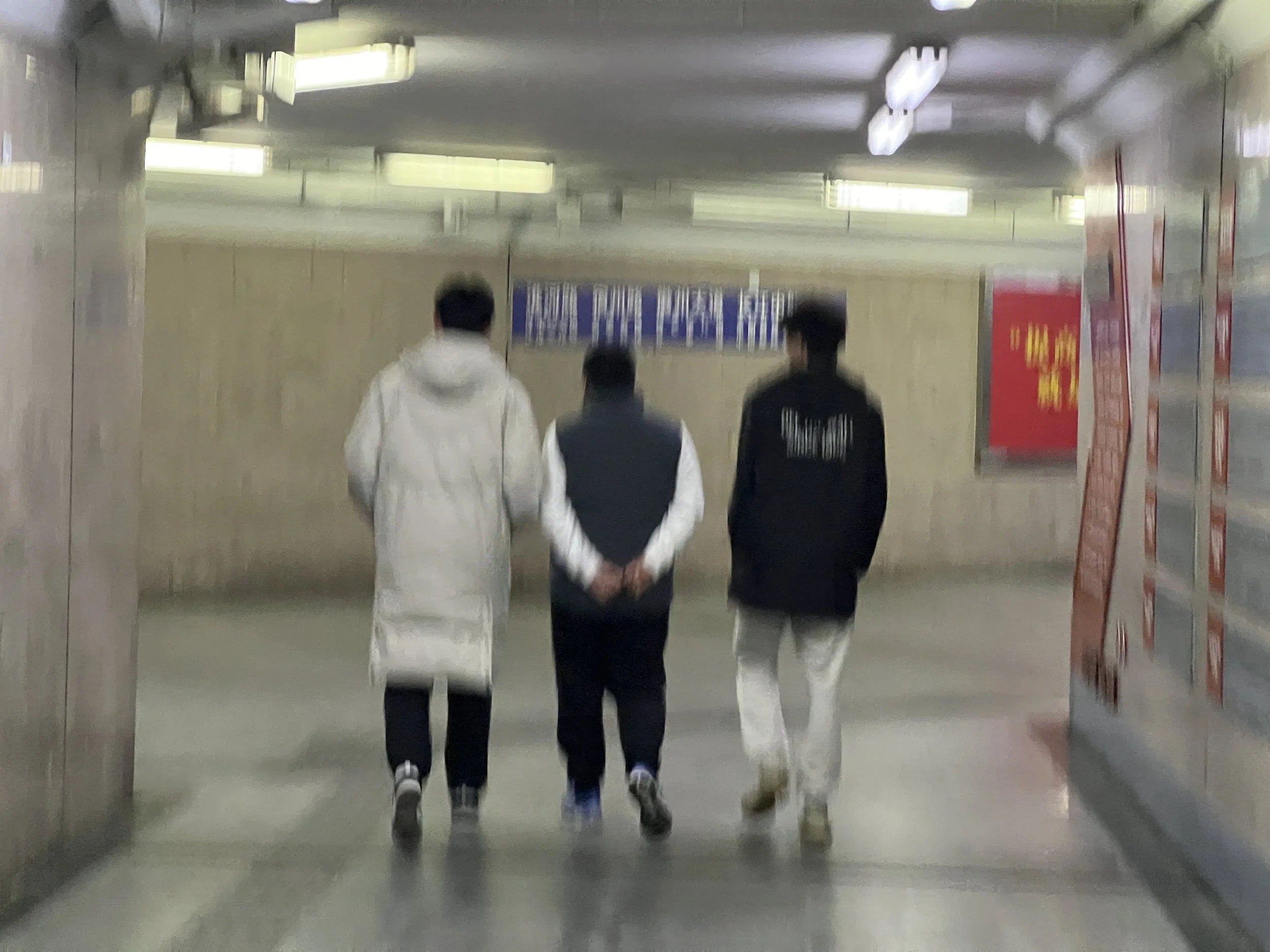 Three people walking through an indoor corridor with bright ceiling lights, two dressed in white and black jackets, and one in a dark hoodie.