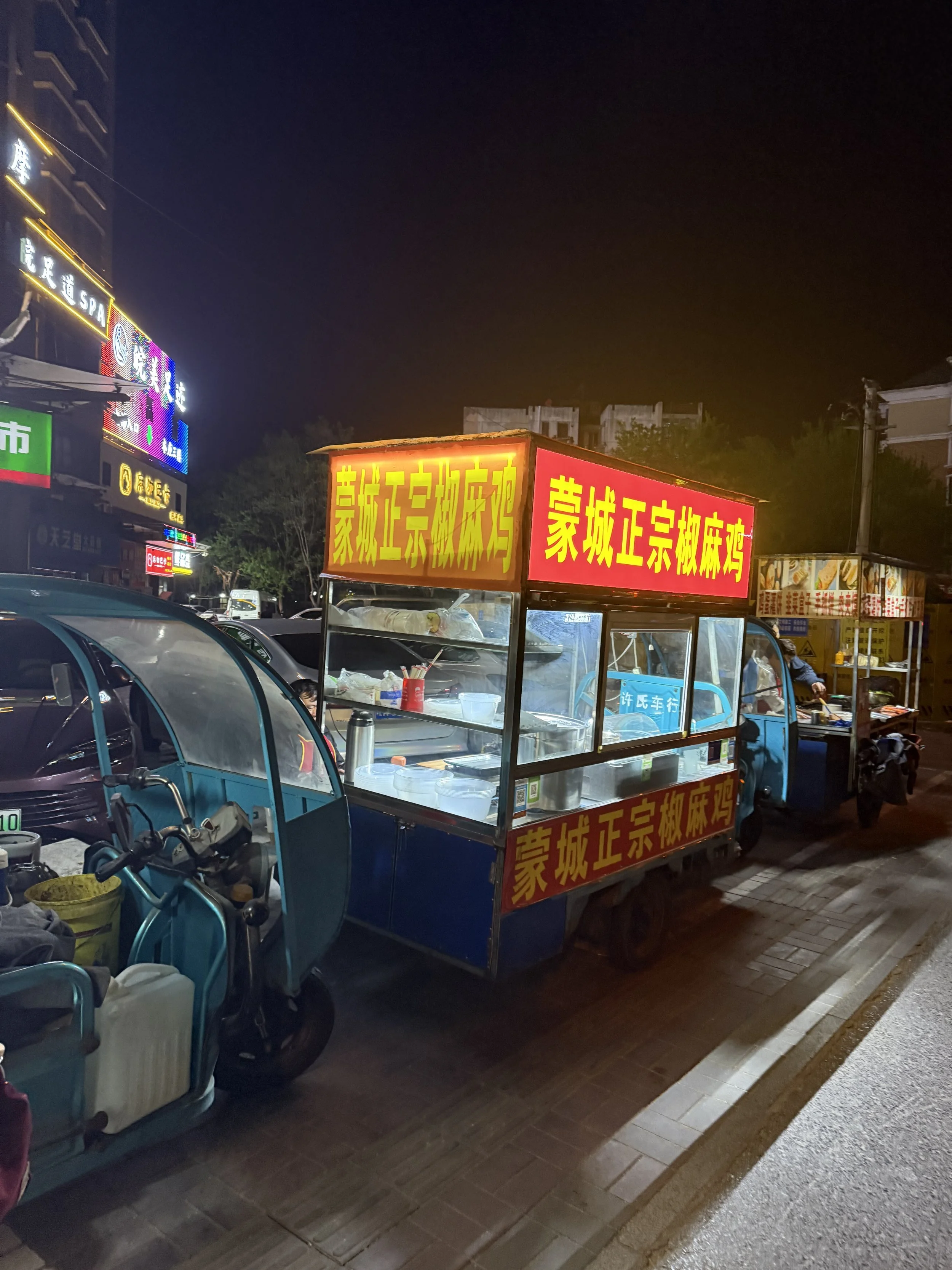 Night scene of street food stalls with bright yellow and red signs in Chinese characters, parked cars, and scooters along a sidewalk.