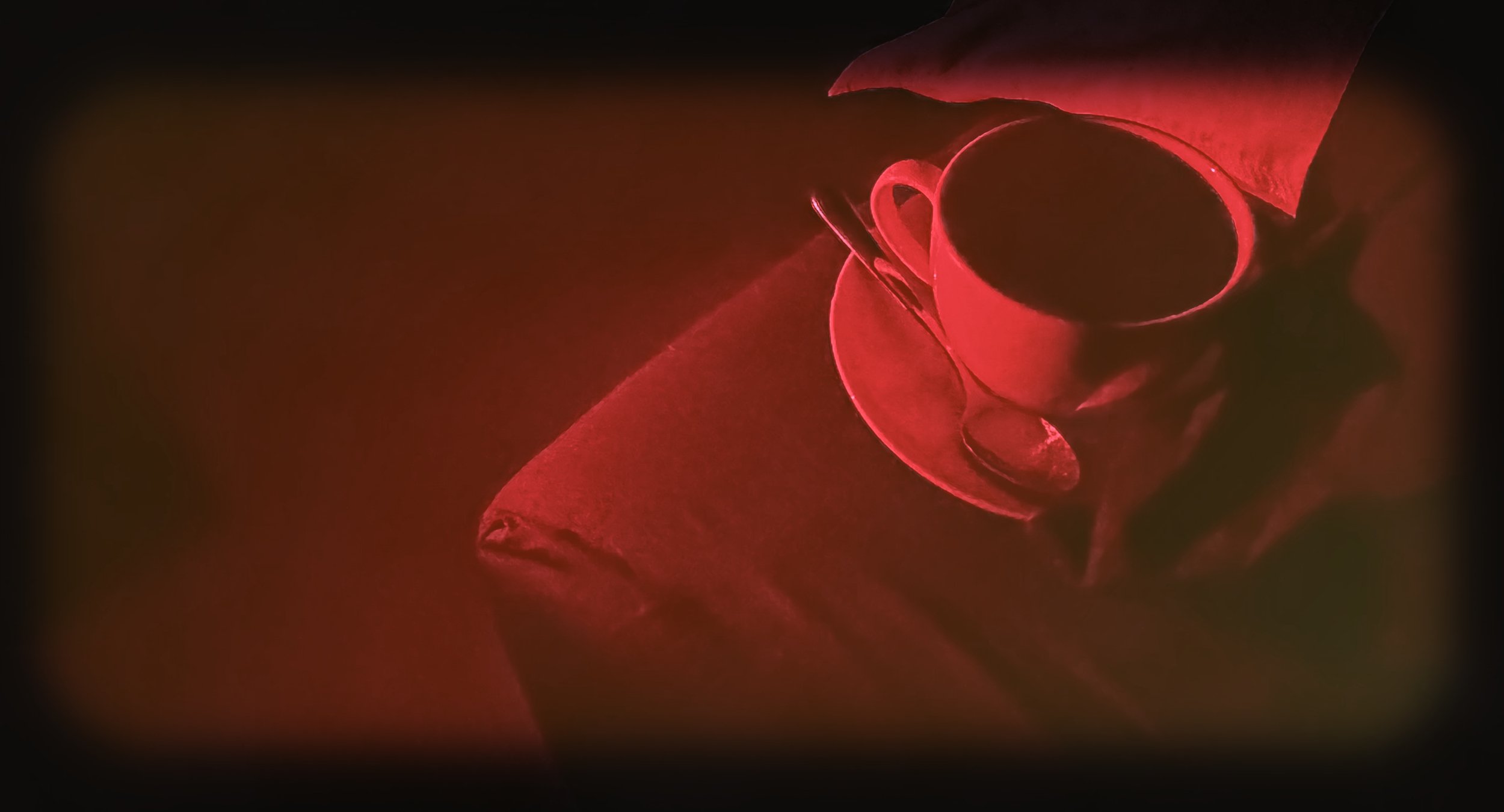 A red-tinted image of a person holding a cup of coffee or tea with a spoon on a white saucer, wrapped in a red cloth or blanket, with a dark background.