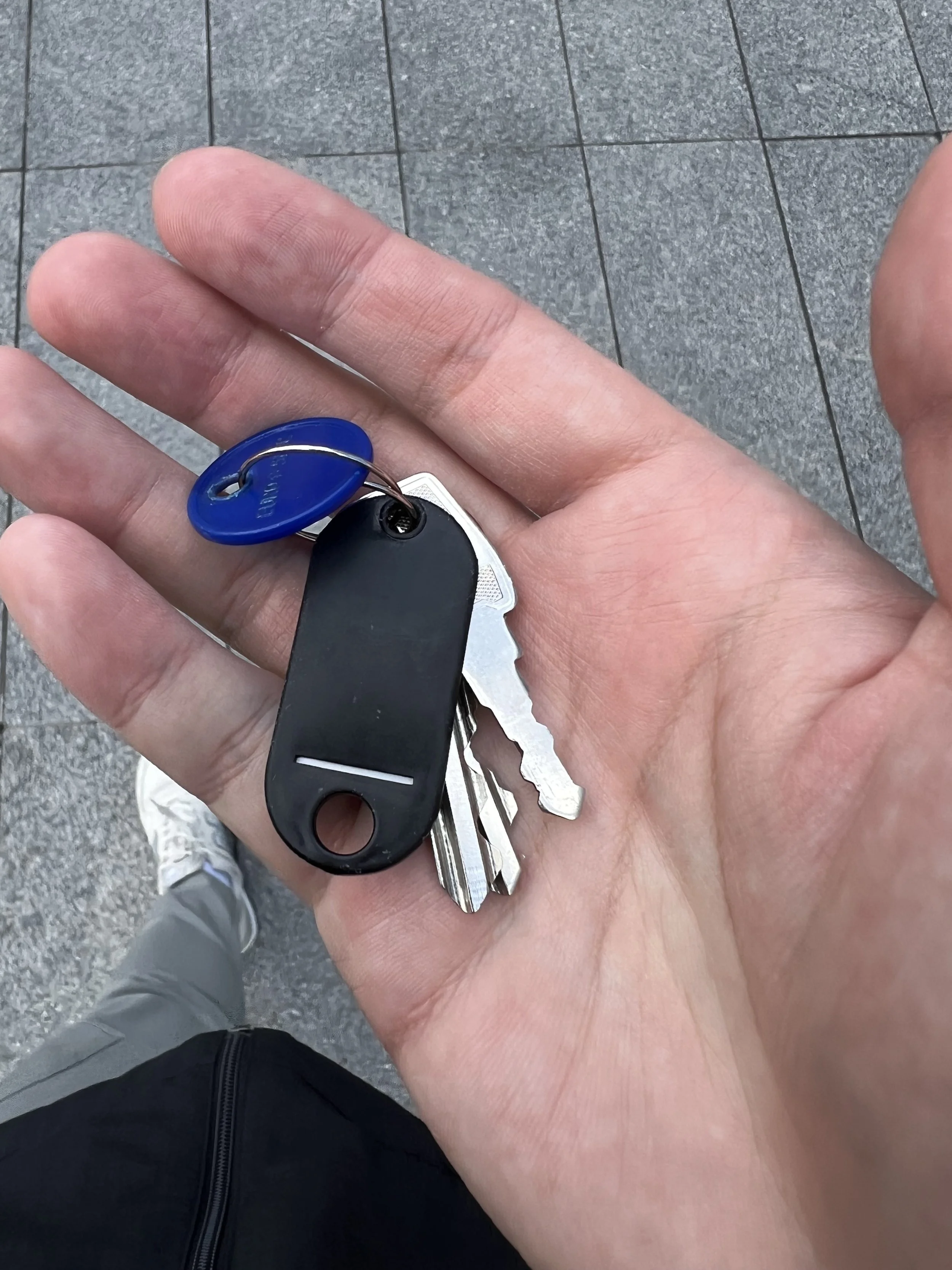 A person's hand holding a set of car keys against a tiled pavement background, with another person wearing gray pants and sneakers visible in the lower part of the image.