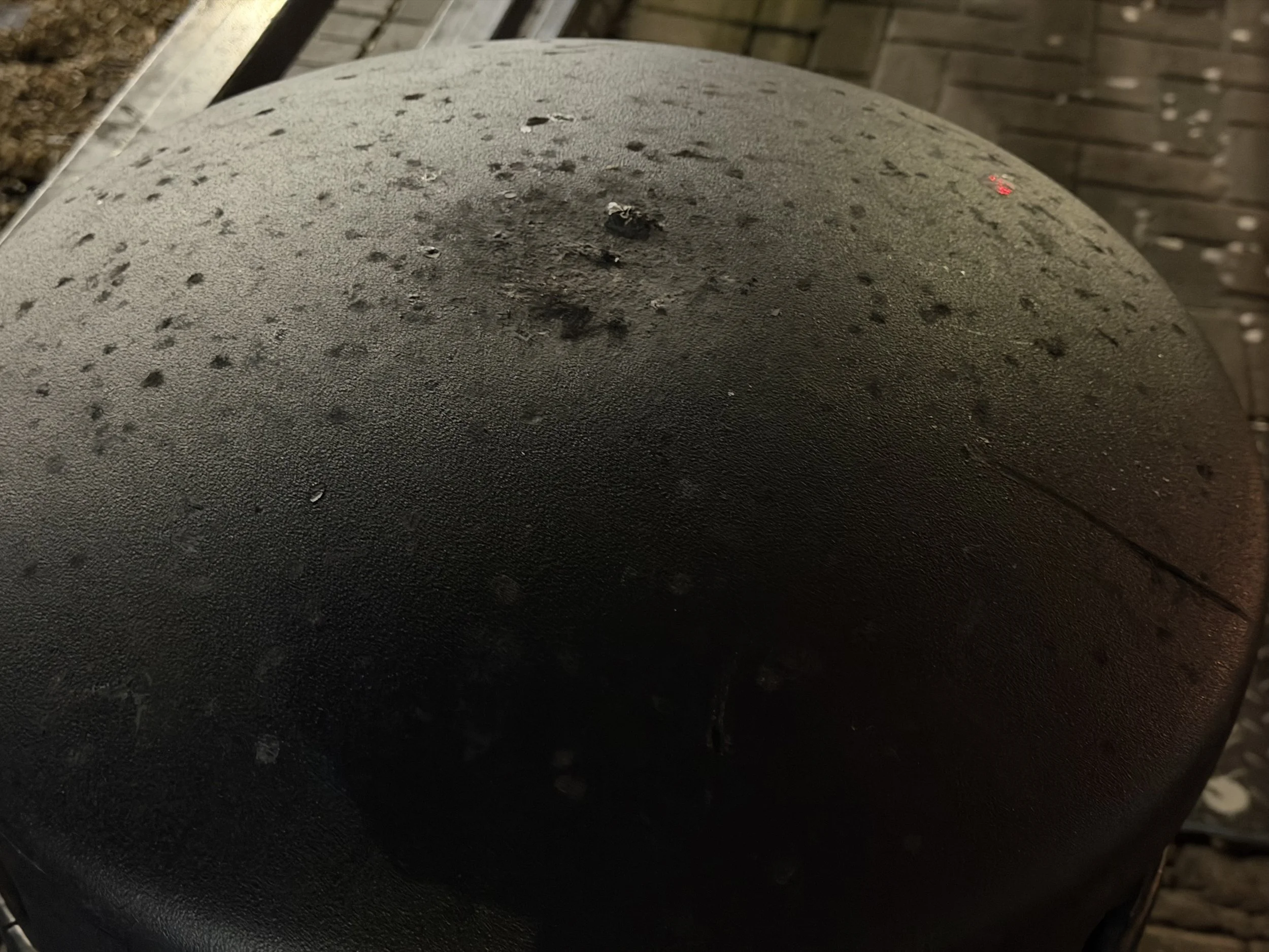 Close-up of a dark, textured surface with small holes and a rough appearance, possibly a tire or rubber object, with a blurred background showing a wooden surface.