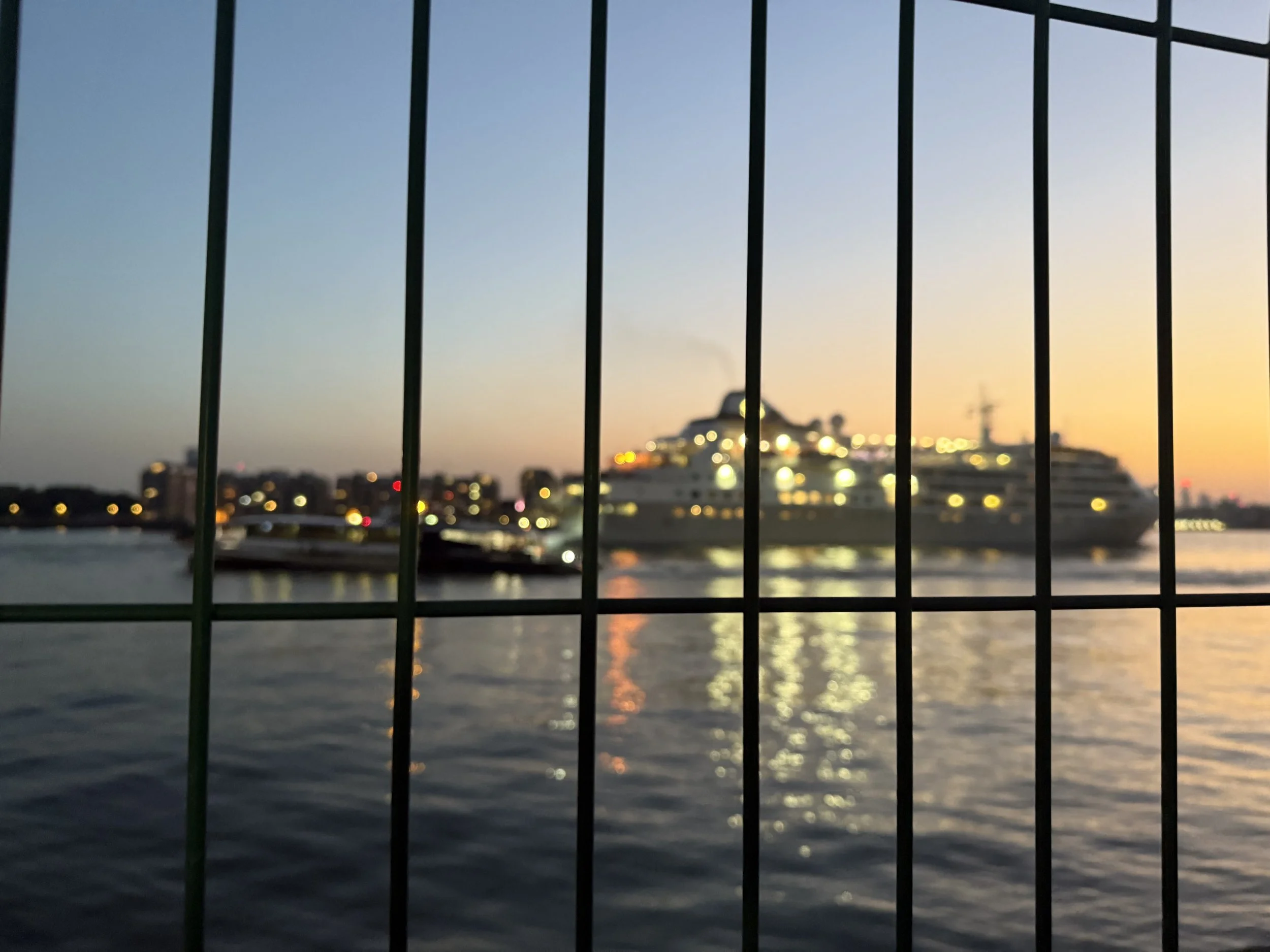 View from a balcony with vertical metal bars overlooking a river, with a boat on the water, a building, and a crane in the distance at sunset with a bright sky.