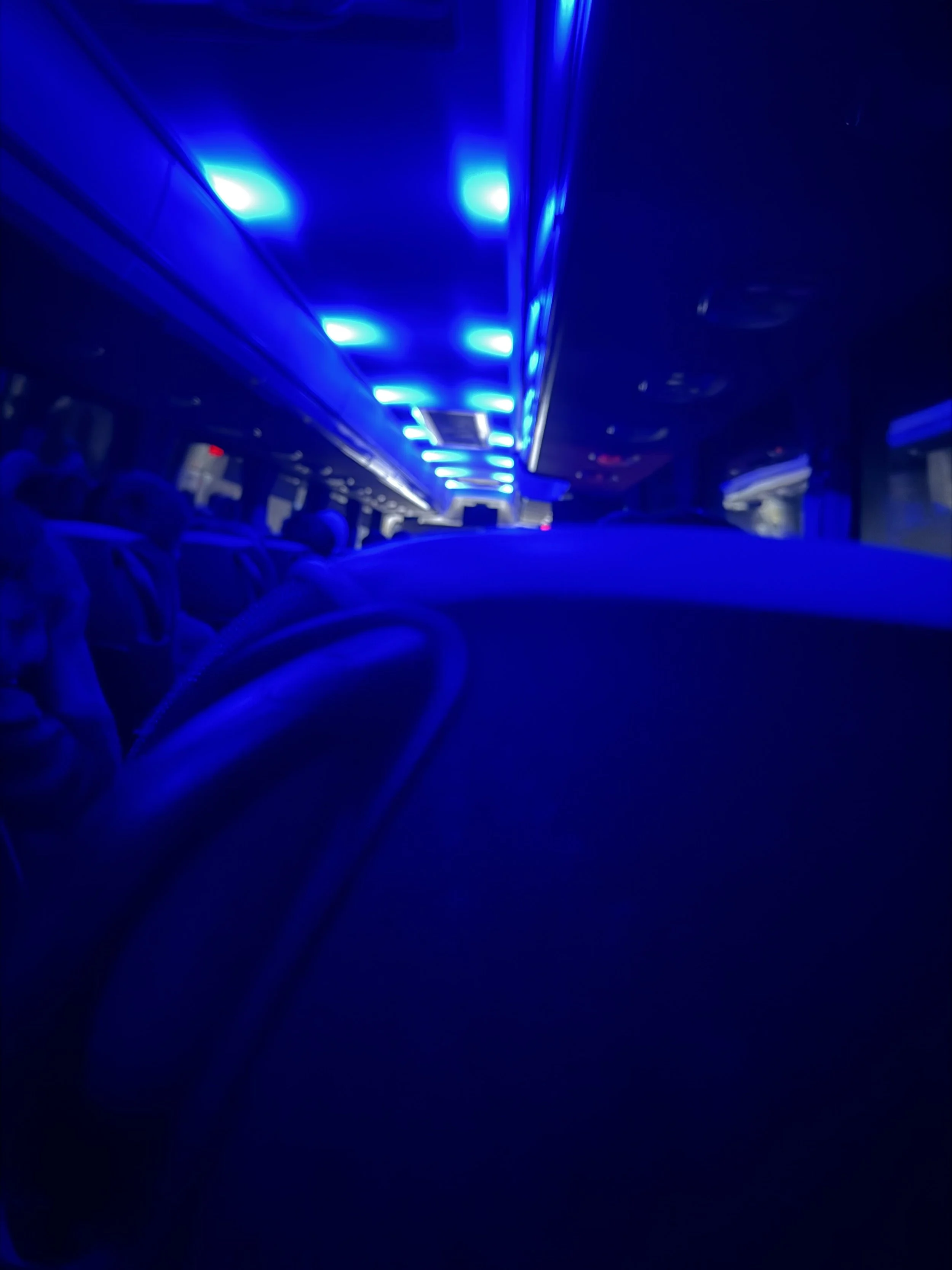 Interior of a vehicle or bus with blue LED lights on the ceiling and some people seated, viewed from the back.