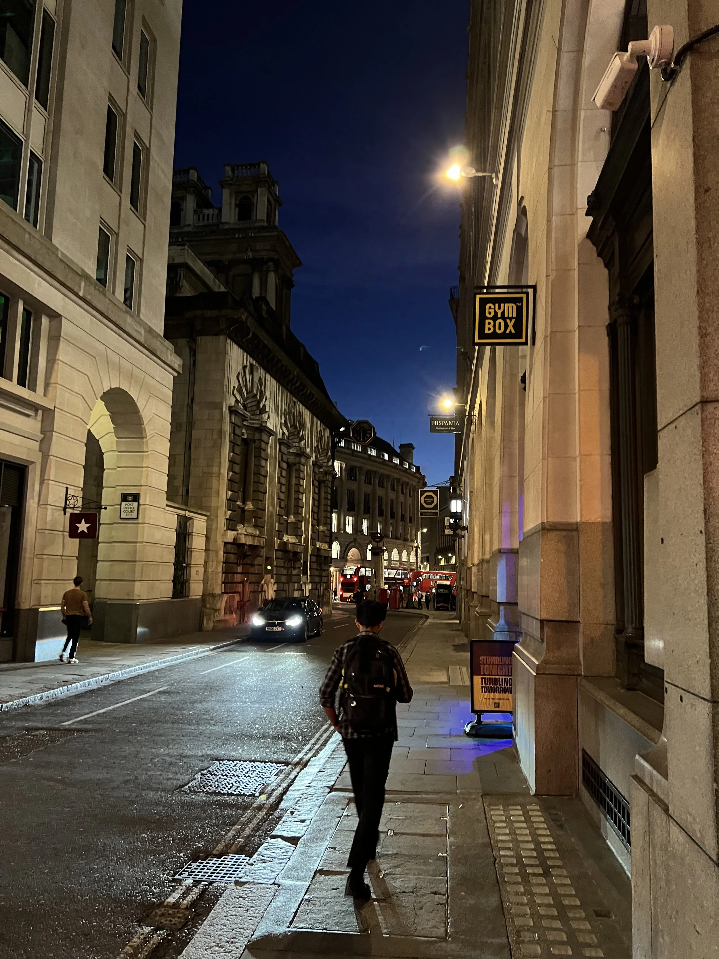 Nighttime street scene with pedestrians, vehicles, and historic buildings, illuminated by streetlights, in an urban area.