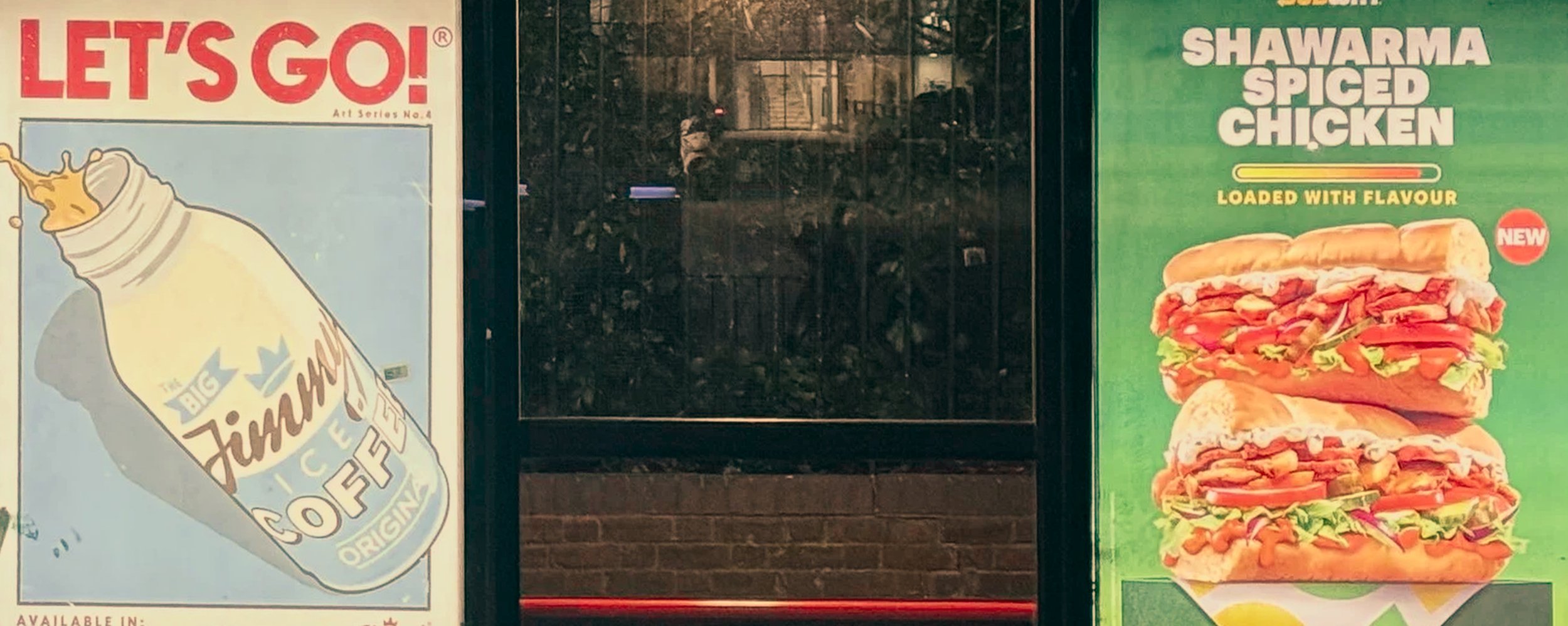 Promotional posters on a storefront window. The left poster is for McDonald's Big Fizziha Coffee with a stylized illustration of a coffee cup. The right poster advertises a Spiced Chicken Shawarma sandwich, showing two large sandwiches filled with ch