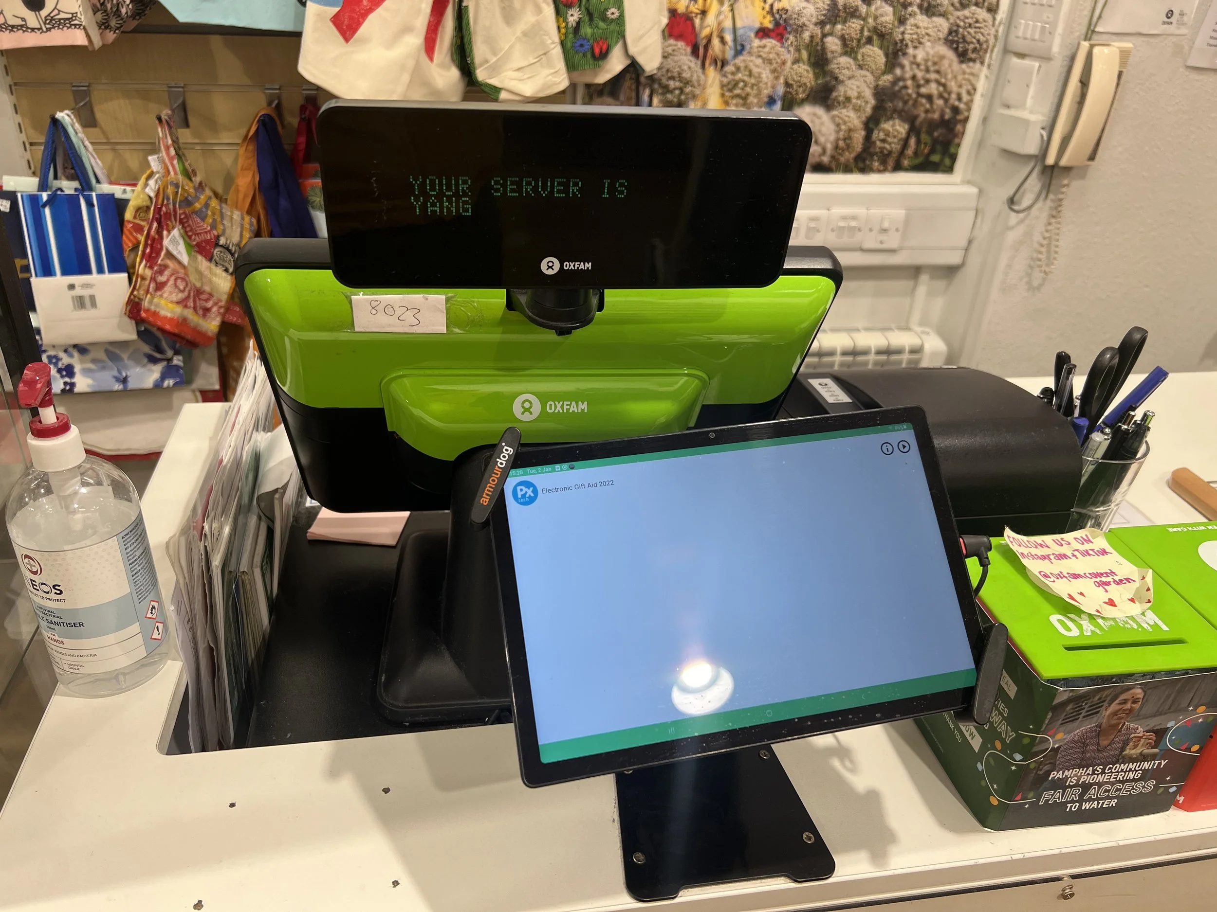 A checkout counter at a store with a green Oxfam donation device displaying the message, "Your server is Yang." There are various items like a water bottle, pens, and colorful fabric bags around the counter, with a display of flowers and other mercha