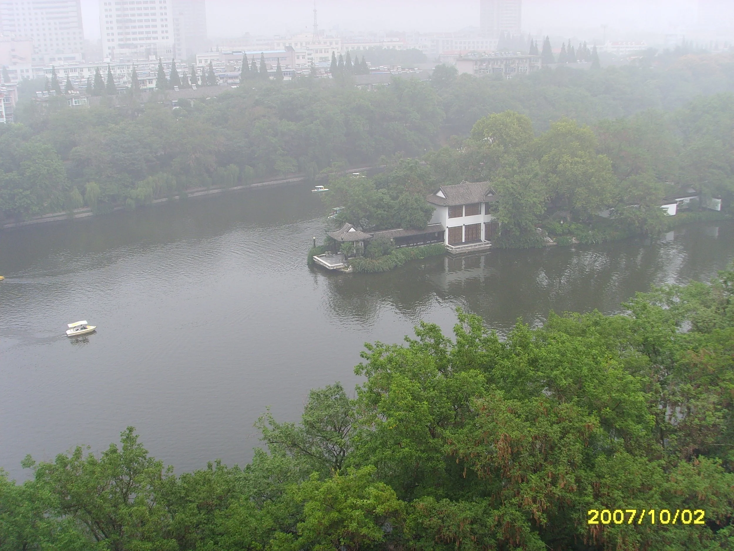 A foggy cityscape with high-rise buildings in the background, a river running through lush green trees, and traditional-style buildings along the water. Two boats are seen on the river, and a timestamp shows the date 2007/10/02.