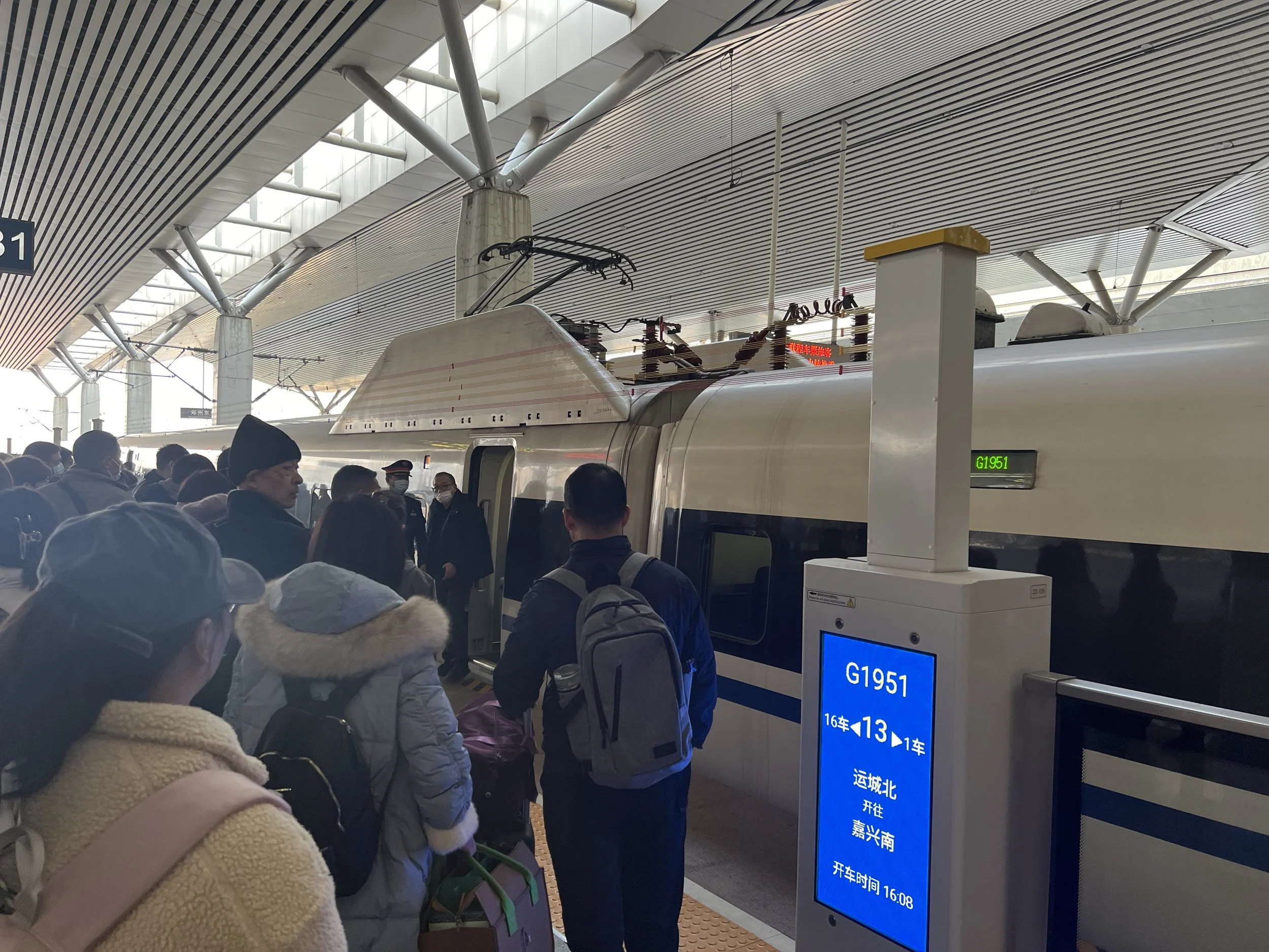 People waiting in line at a train station platform, with a high-speed train in the background, digital sign showing train information, and modern station architecture.