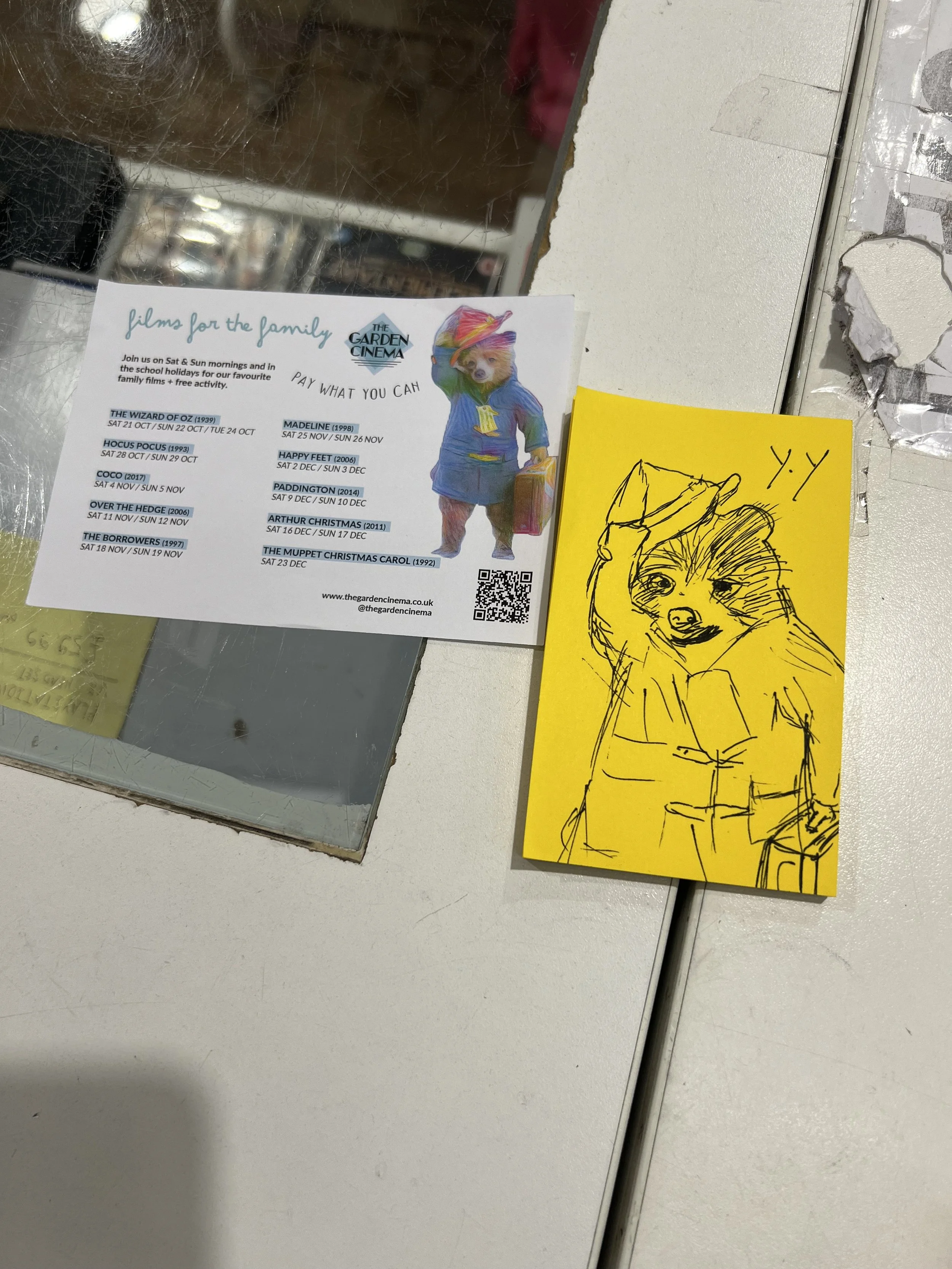 A mirror reflecting the image of a white and yellow flyer and a yellow drawing paper on a table surface. The flyer contains information about a film screening event, with a picture of a bear dressed as a human and a list of film titles and dates. The
