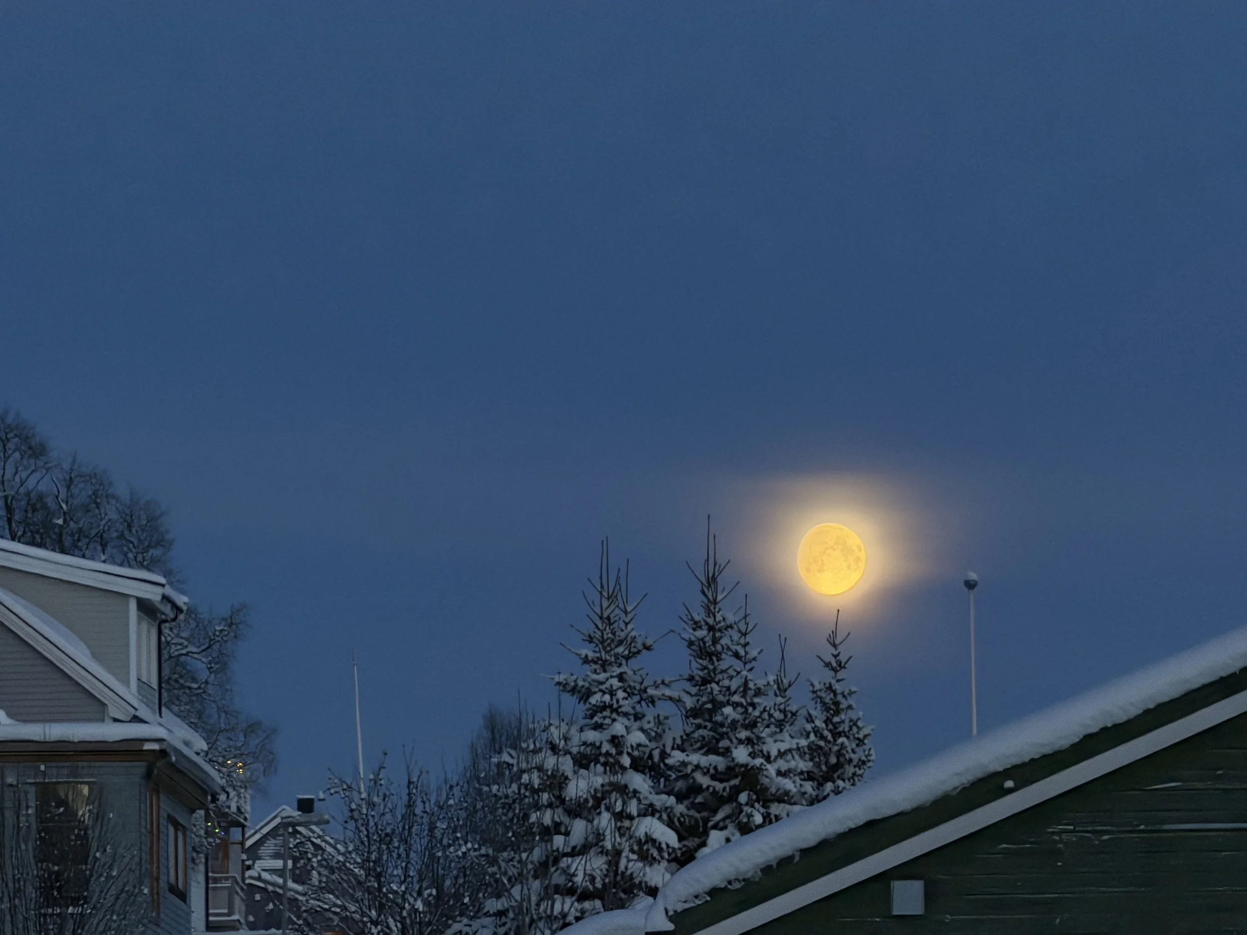 A nighttime scene of a clear blue sky with a bright full moon near the horizon, snow-covered rooftops, leafless trees, and some house windows visible in the foreground.