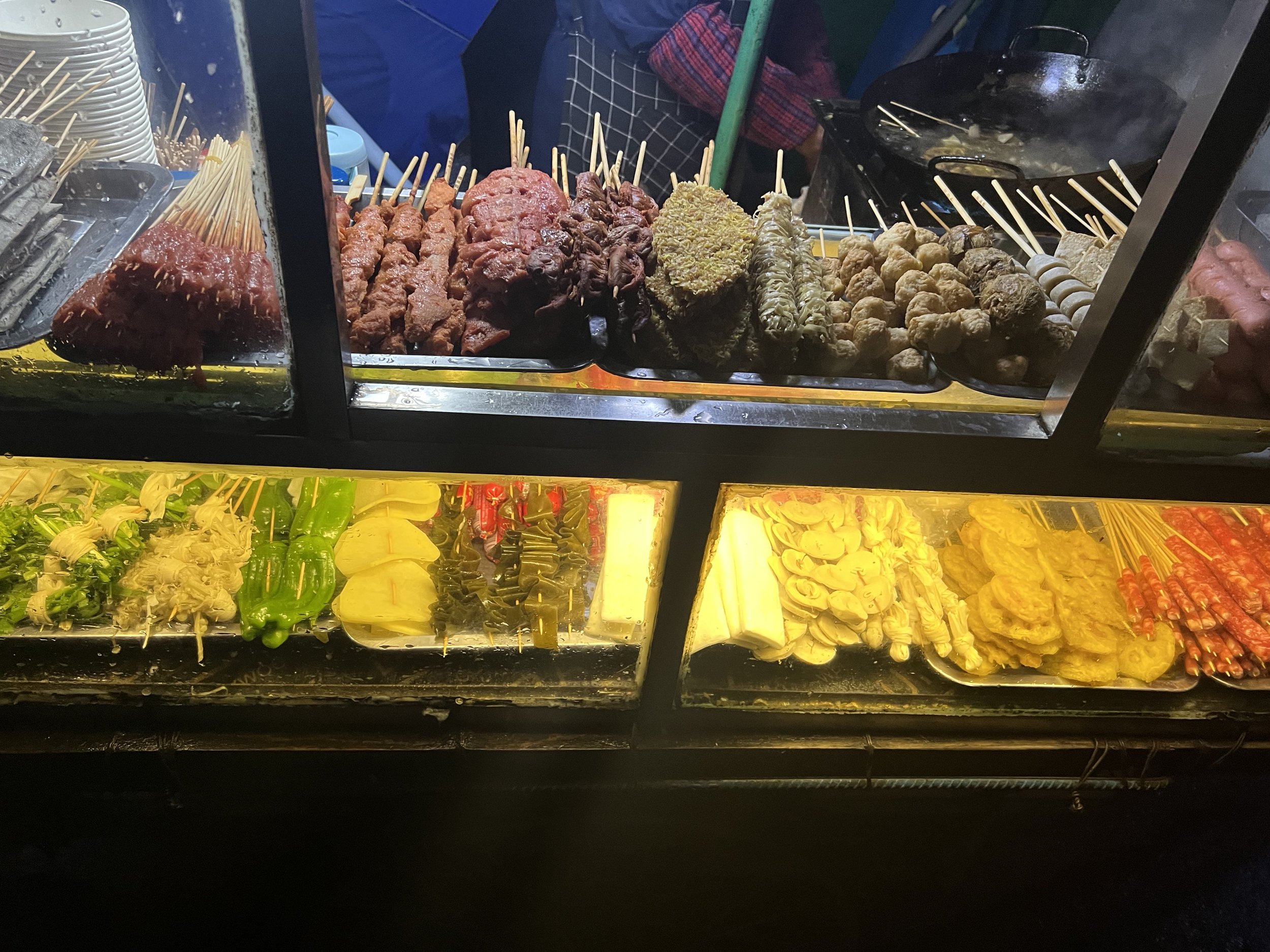 Street food stall with skewered meat and vegetables, some cooking in a hot pan behind a glass display case.