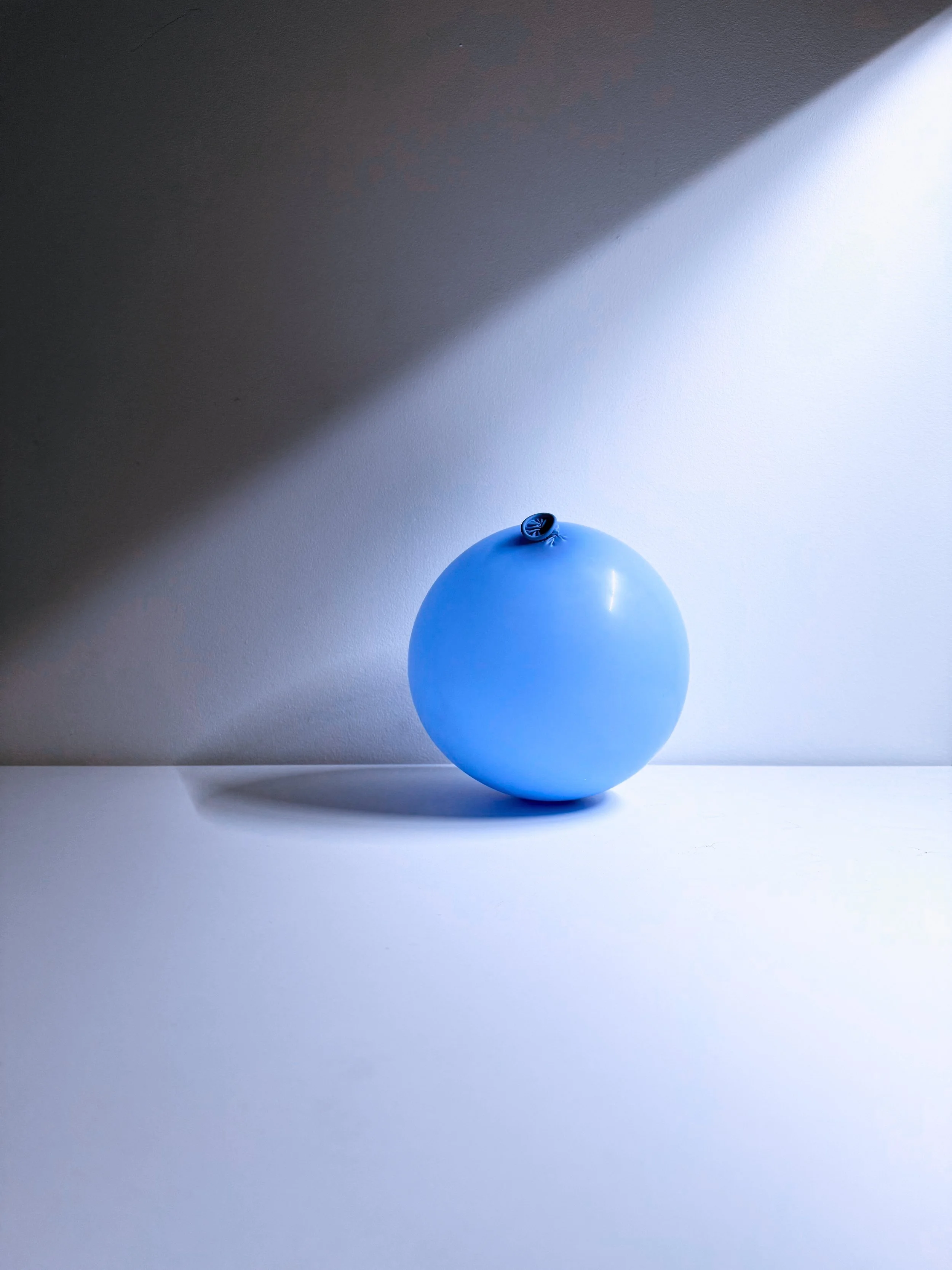 A blue balloon on a white surface with a shadow cast on the wall behind it, illuminated by natural light.
