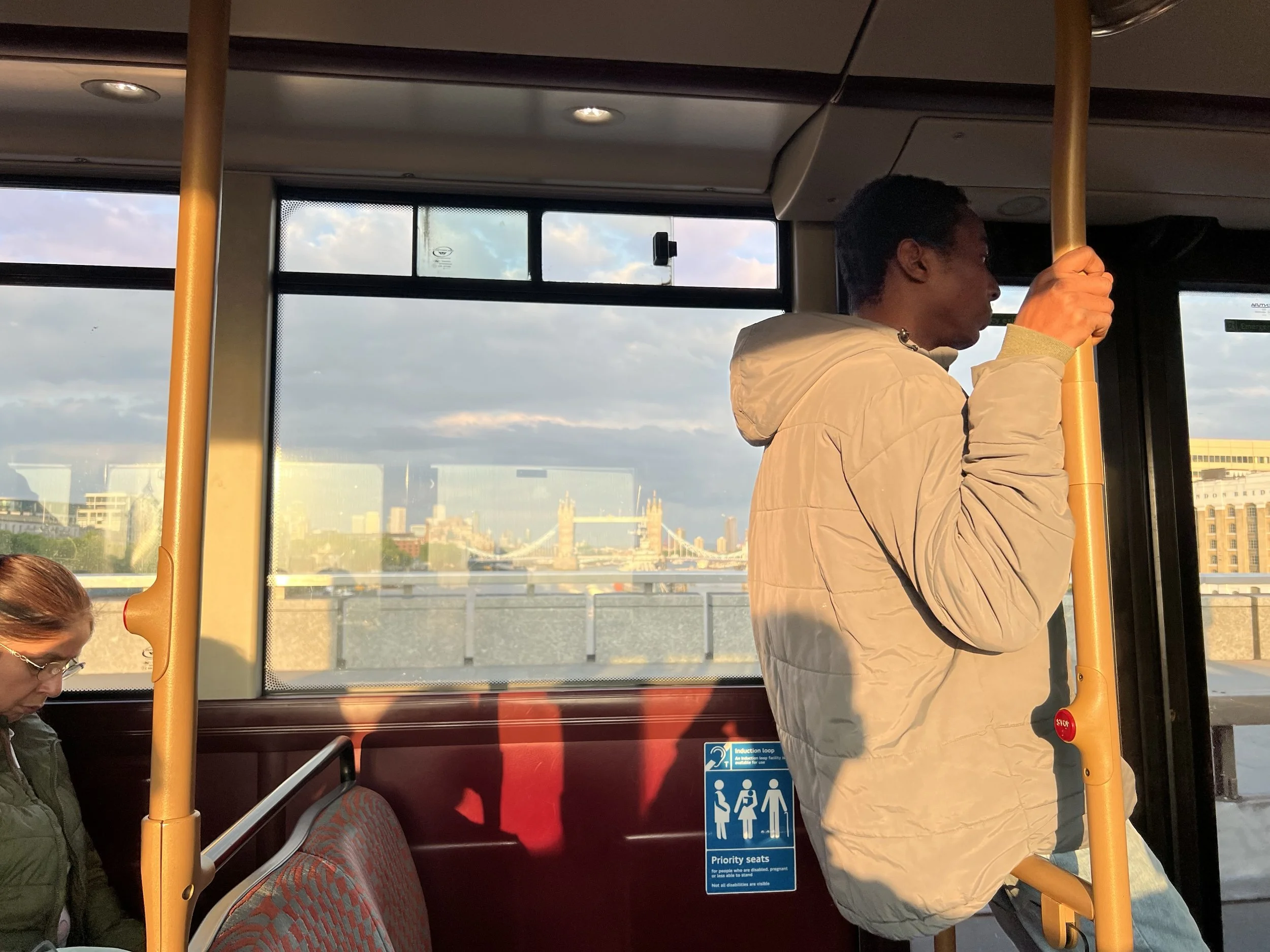 A woman in a beige jacket standing, holding onto a yellow pole inside a bus, with a city skyline and cloudy sky visible through the windows.