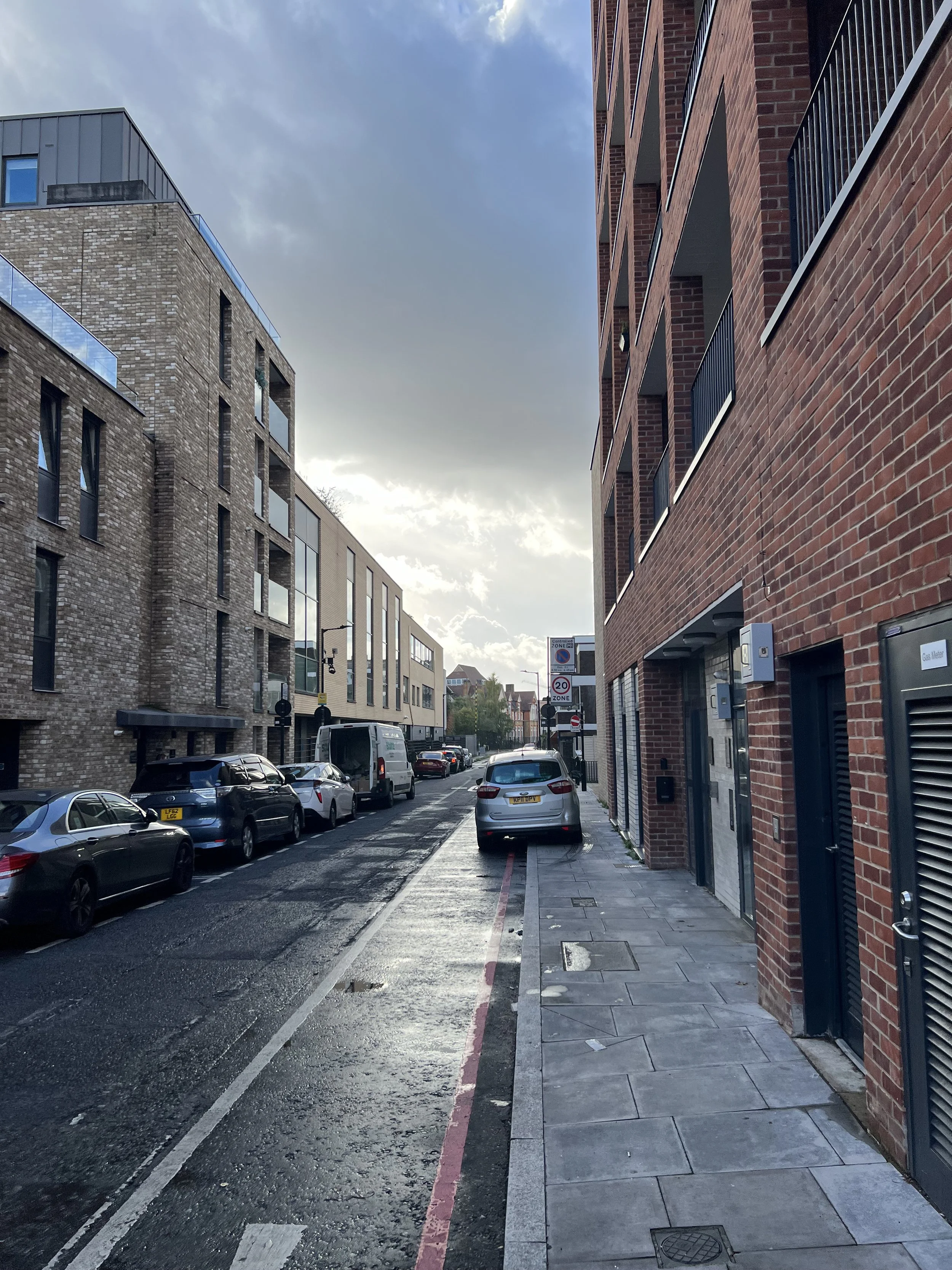 A city street scene with parked cars along the curb, modern brick and glass buildings on either side, and a cloudy sky overhead.