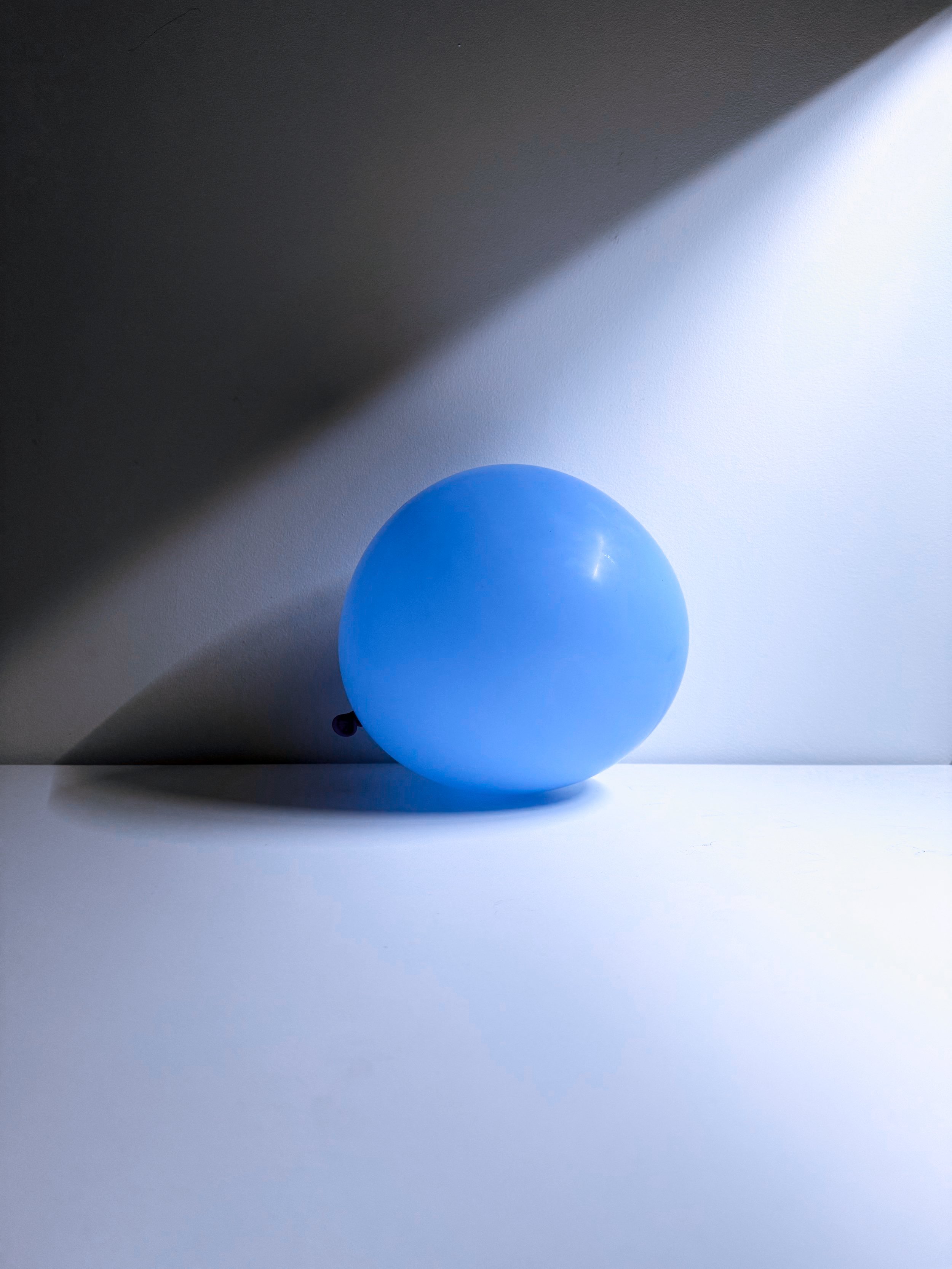 A blue balloon on a plain white surface with shadows cast by light coming from the upper left.