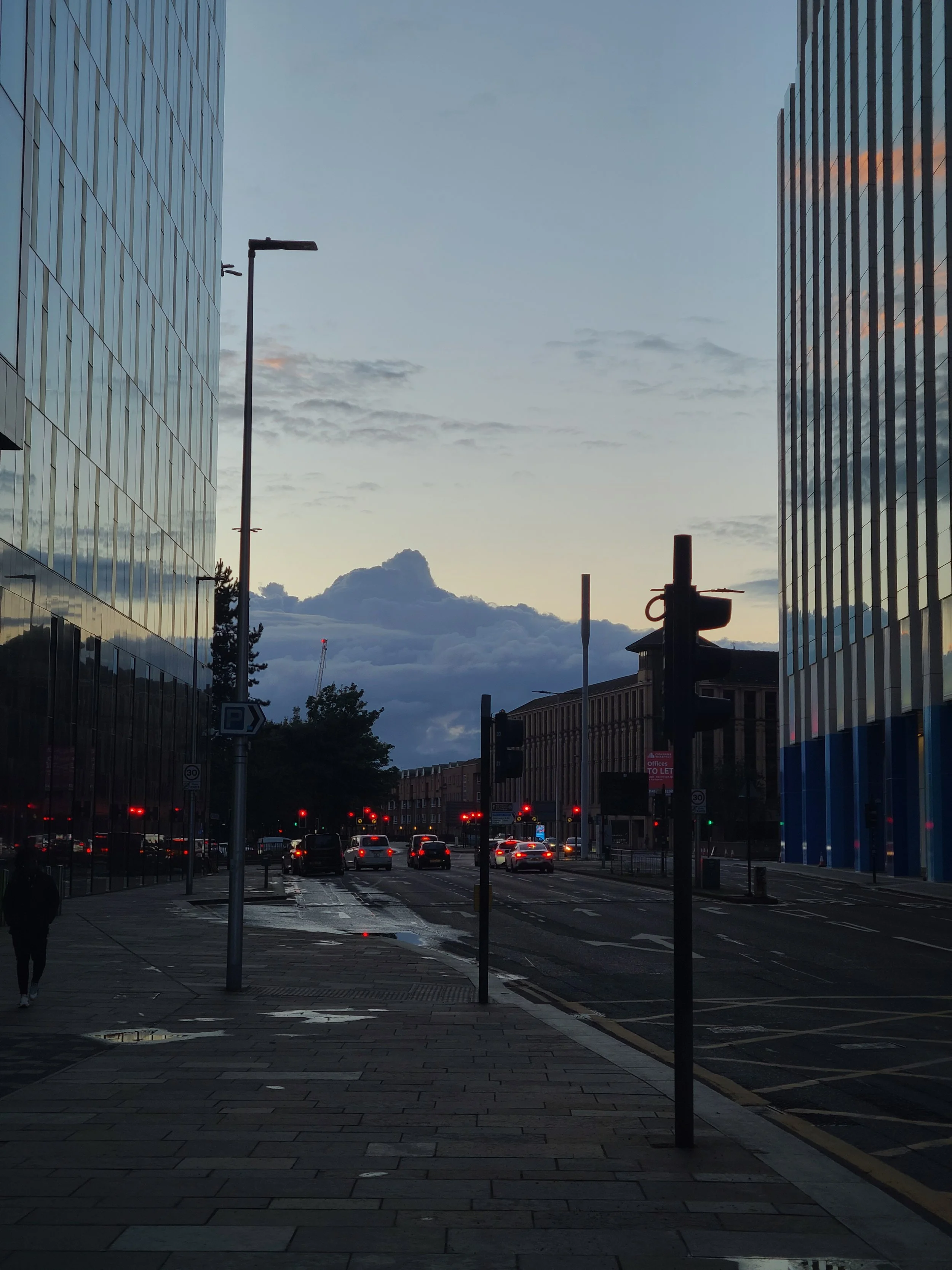 City street scene during dusk with modern office buildings on both sides, traffic lights showing red, several cars on the road, and a cloudy sky with a mix of dark and light clouds.