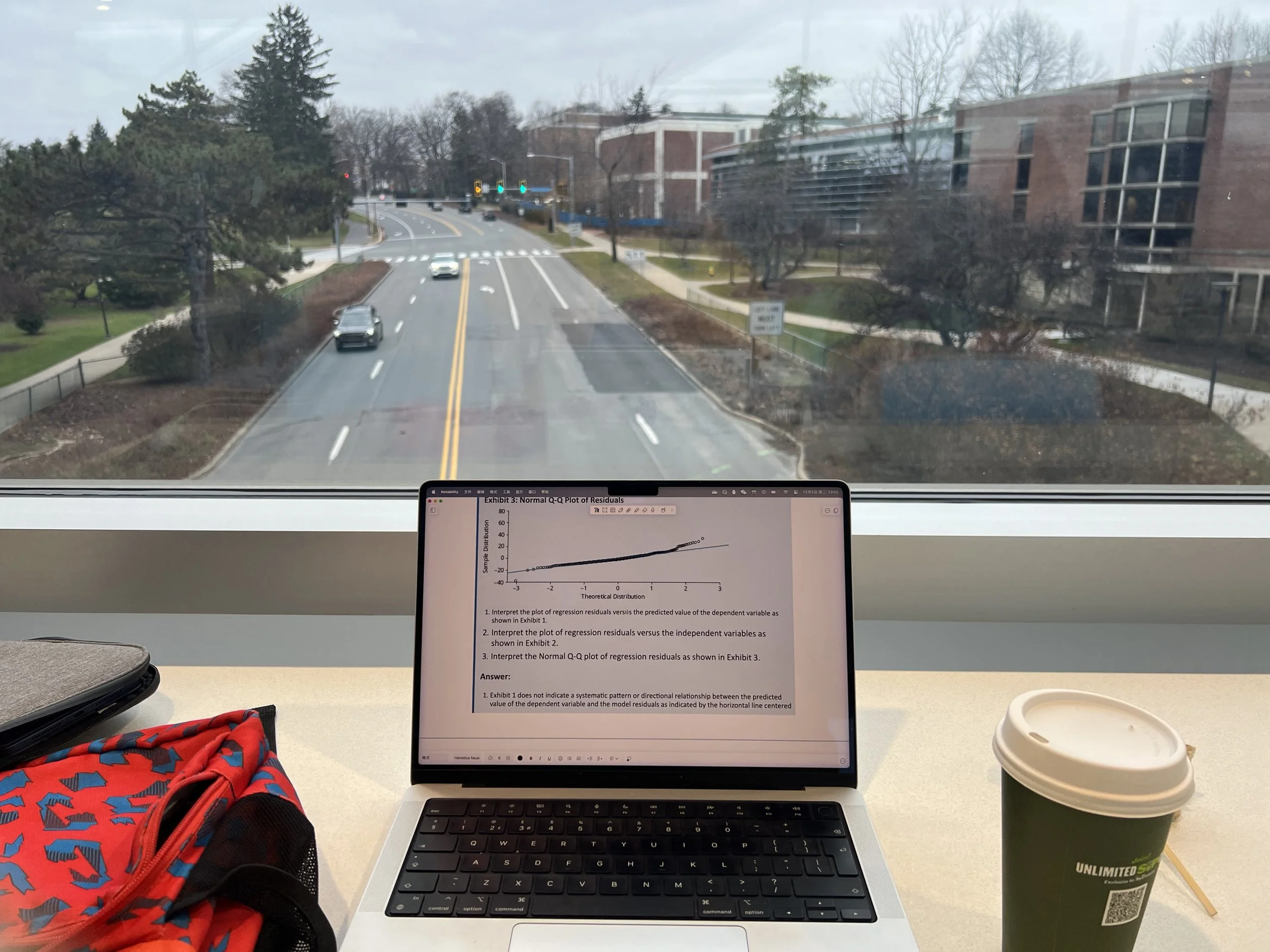 A view from inside a building showing a desk with a laptop displaying a scientific graph, a red backpack with blue and black patterns, a disposable coffee cup, and a window looking out onto a street with trees and buildings.