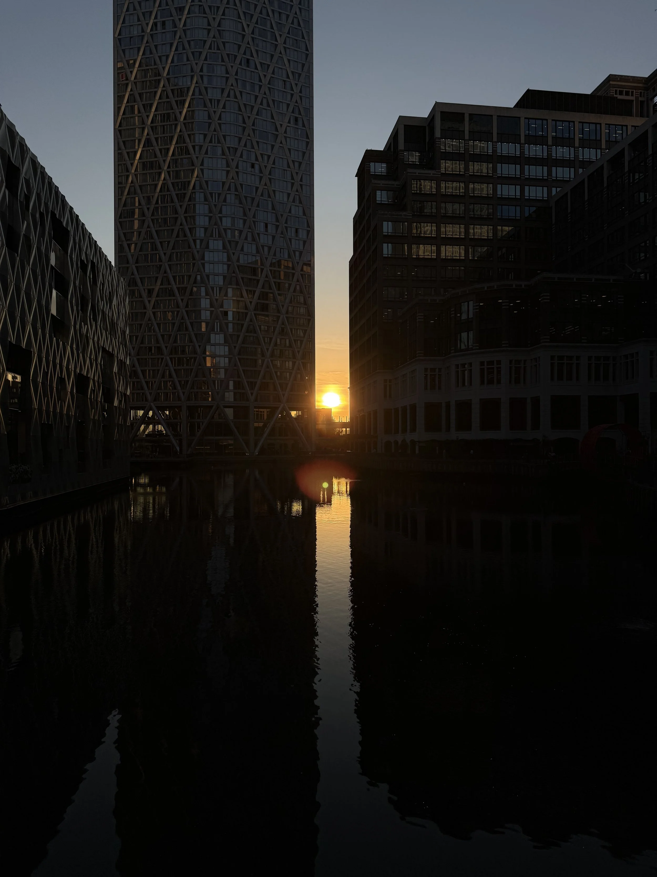 A cityscape at sunset featuring modern high-rise buildings reflected in a waterway with a bright sun near the horizon.