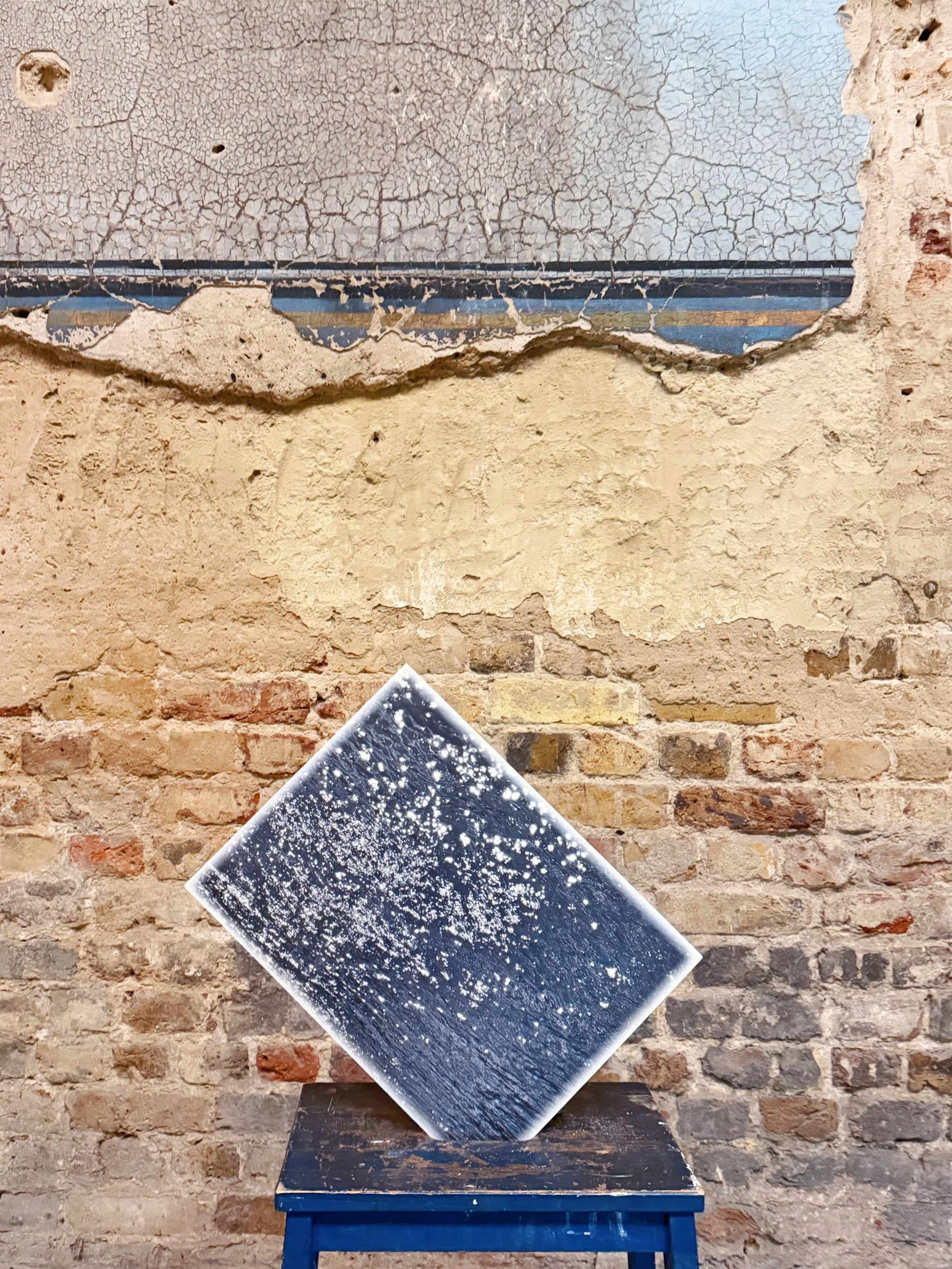 An art installation features a black and white starry sky photograph mounted at an angle on a rustic brick wall, with a large crack running horizontally across the top portion of the wall. The photograph is resting on a dark blue wooden table.