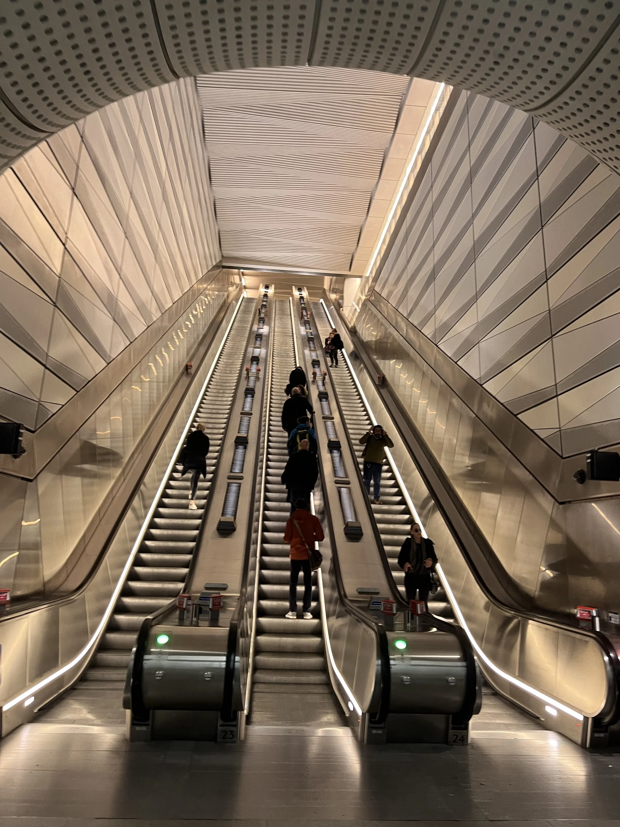 Photo of multiple escalators in a modern indoor setting, with several people ascending and descending. The walls are decorated with sleek, geometric patterns and the ceiling has a contemporary design with lines and dots.