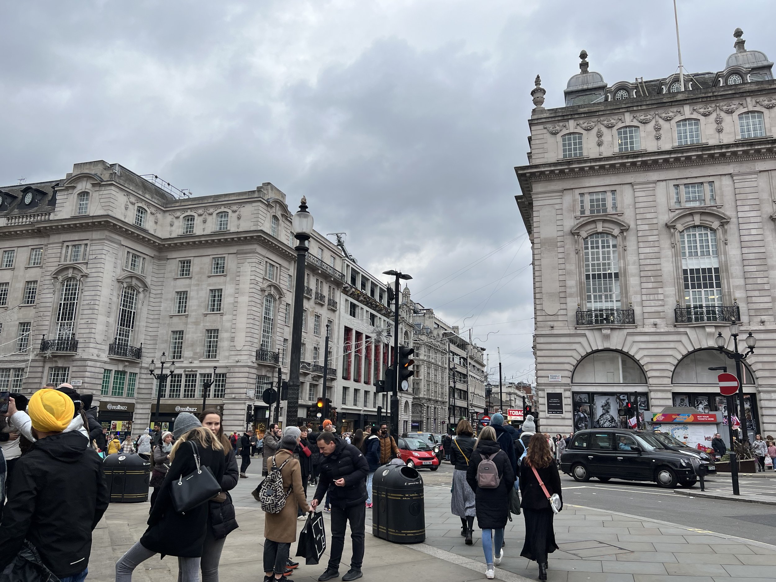 Crowded street in a city with historic buildings, streetlights, and traffic signals under cloudy sky.